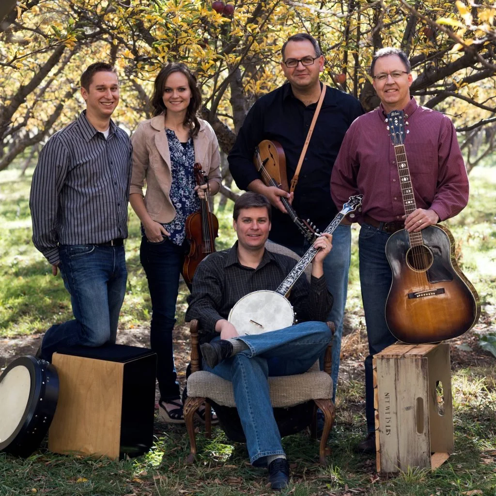 A group of five people with musical instruments in an orchard with yellowing autumn leaves.