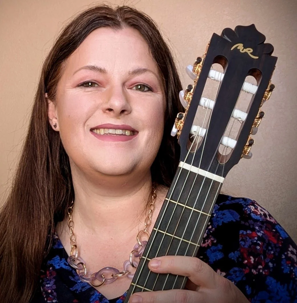 A woman with long brown hair, smiling, holding a black Classical acoustic guitar with white tuning pegs and gold accents.
