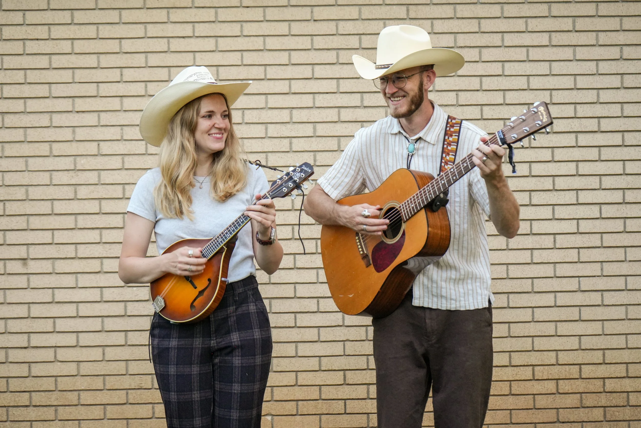 A man and woman wearing cowboy hats playing guitars, smiling and looking at each other against a brick wall.