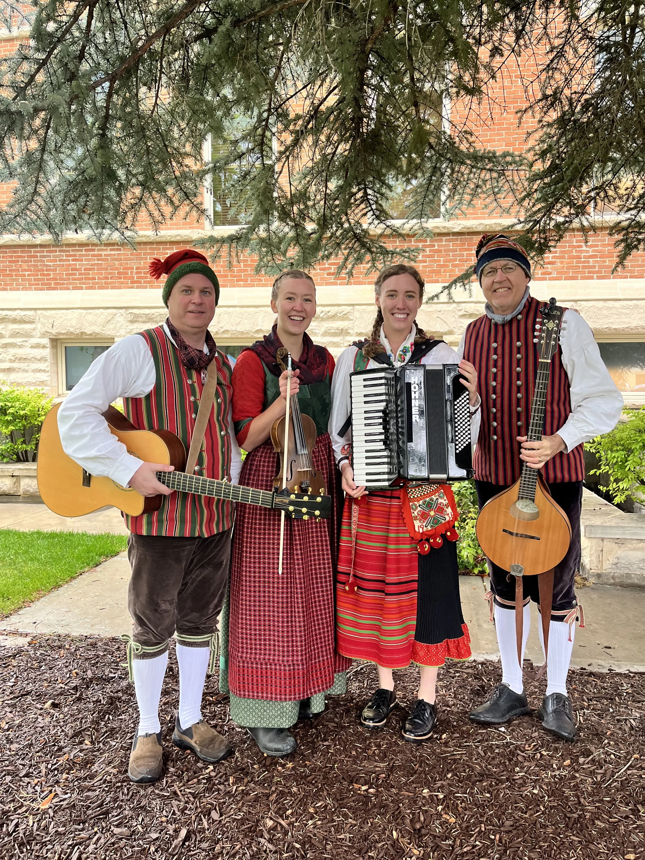 Group of four people dressed in traditional German outfits, holding musical instruments, standing outdoors under a large tree with a red brick building in the background.