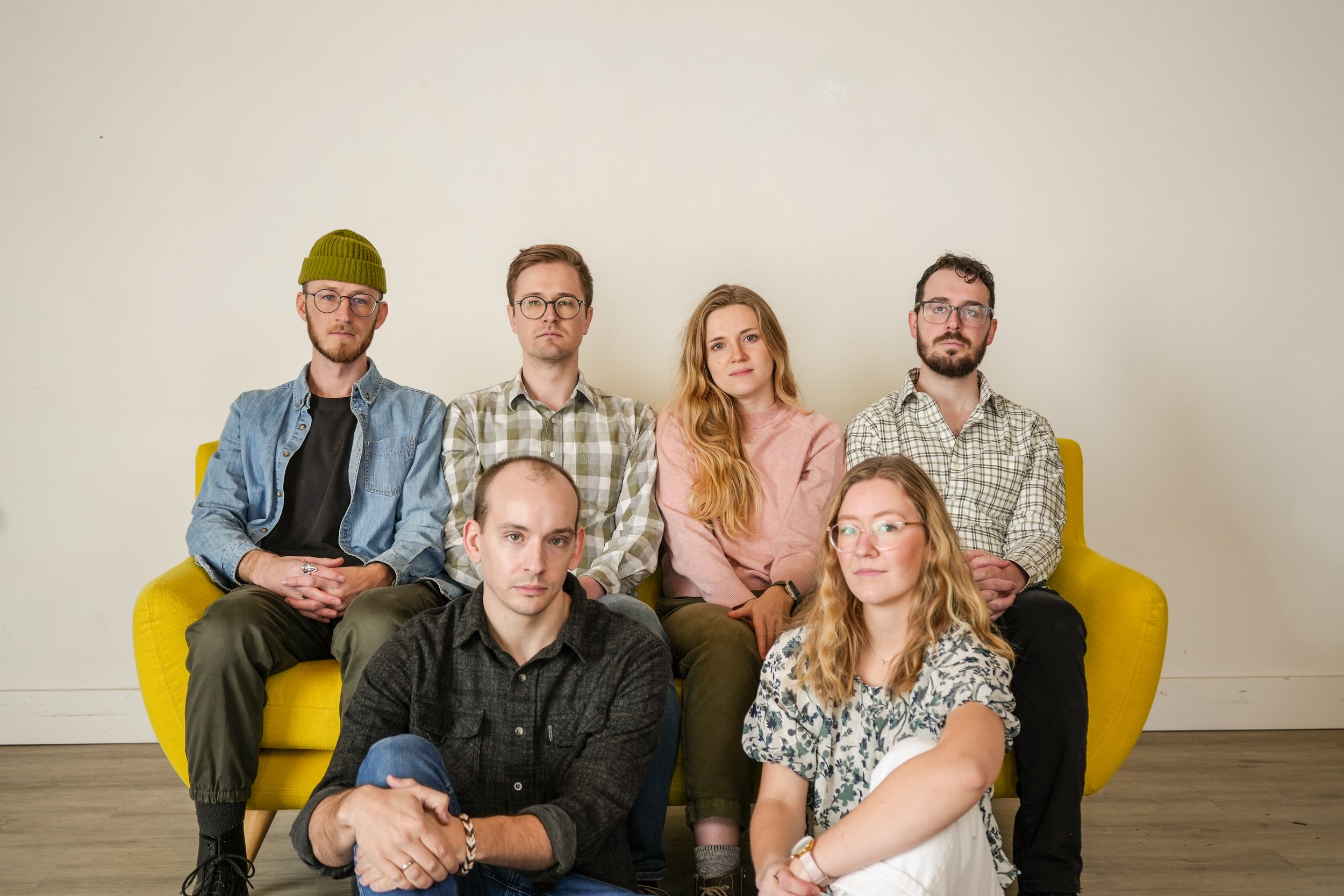 A group of eight young adults sitting on a yellow sofa against a plain white wall, with three women and five men. They have casual attire and serious expressions.