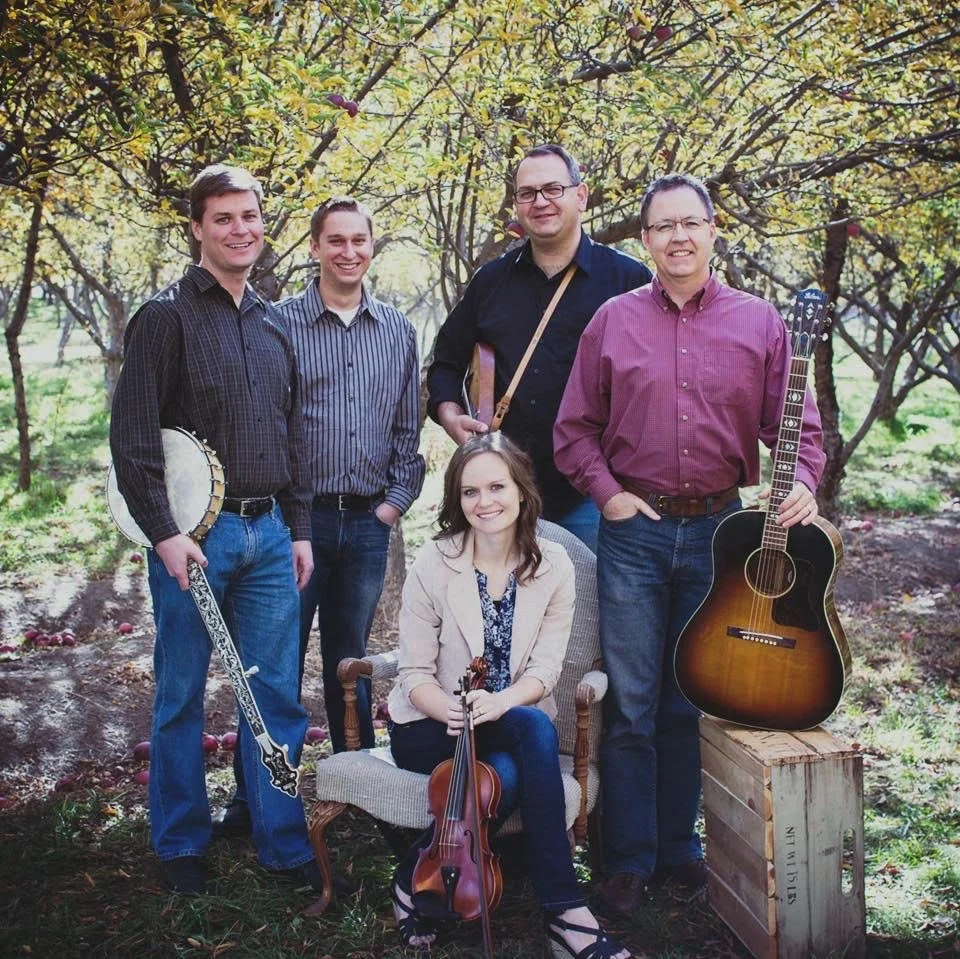 A group of five musicians outdoors surrounded by trees, with one sitting on a chair holding a violin, and four standing with one holding a banjo, one holding a guitar, and one with a musical instrument strap. They are smiling and dressed casually.