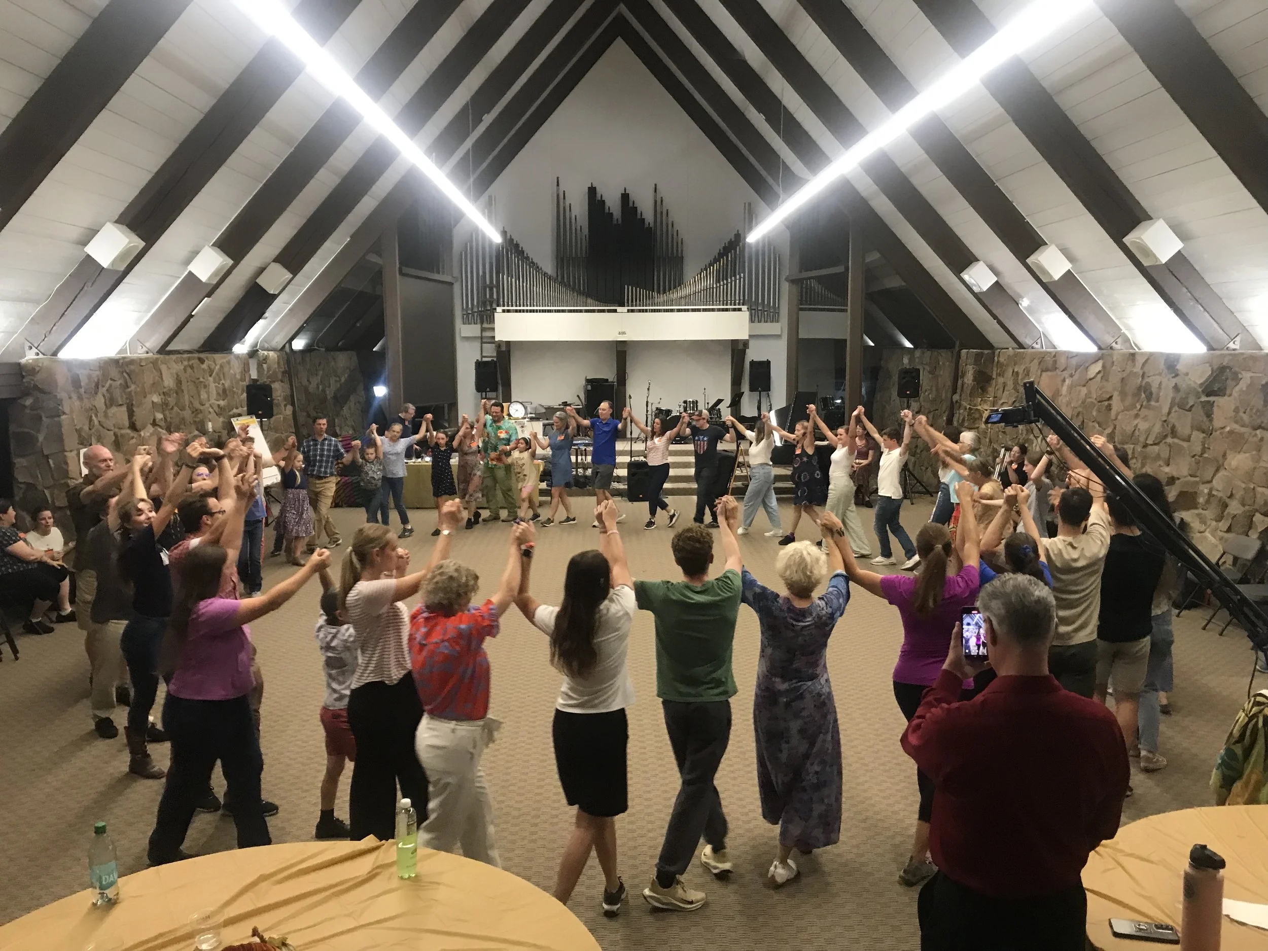 People holding hands and dancing in a circle in a large indoor hall with stone walls and a high, peaked ceiling. A stage with musical instruments is in the background.