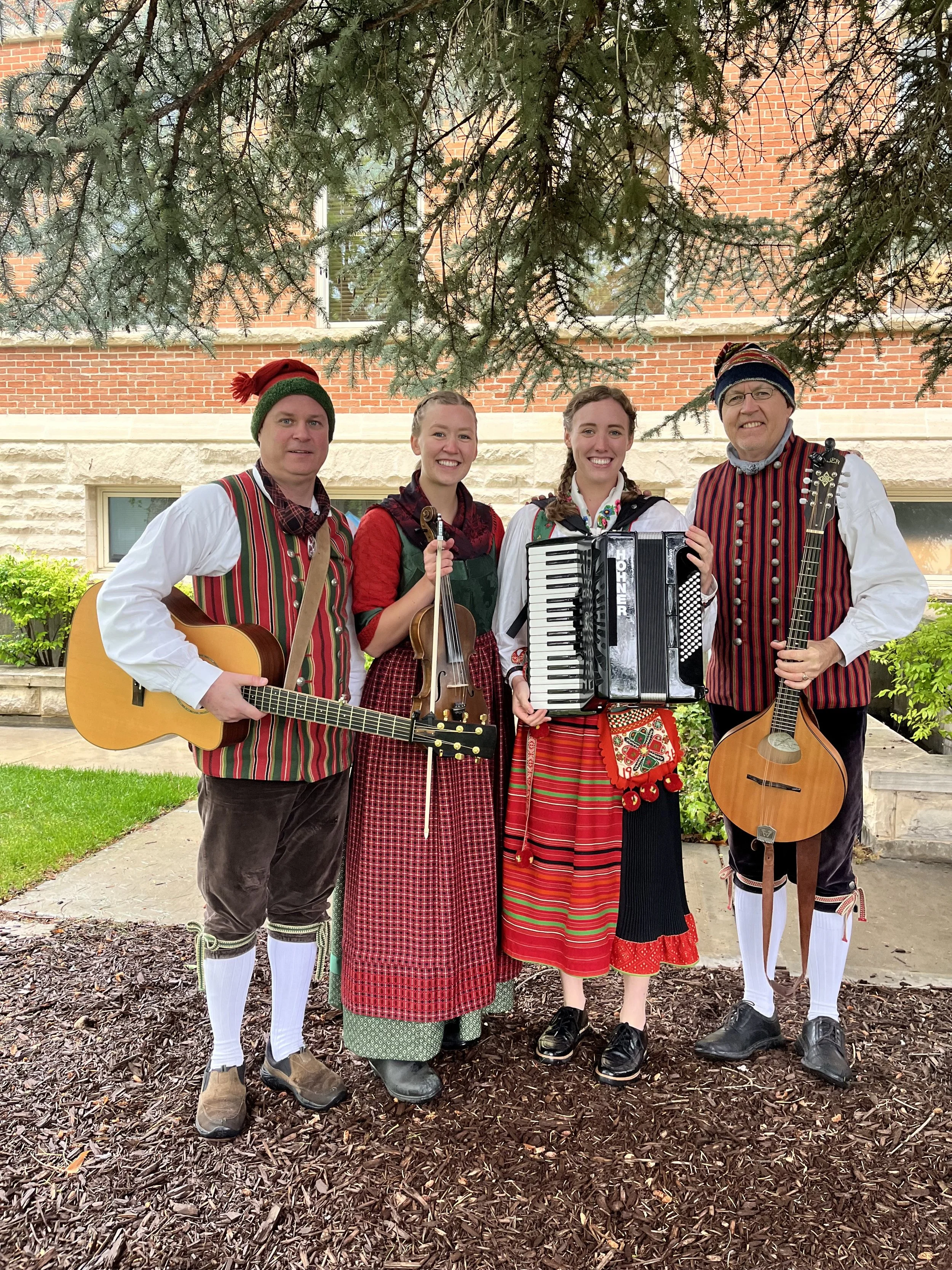 Four people dressed in traditional Scandinavian clothing standing outdoors under a large tree, holding musical instruments such as guitars, a violin, and an accordion, with a red brick building in the background.
