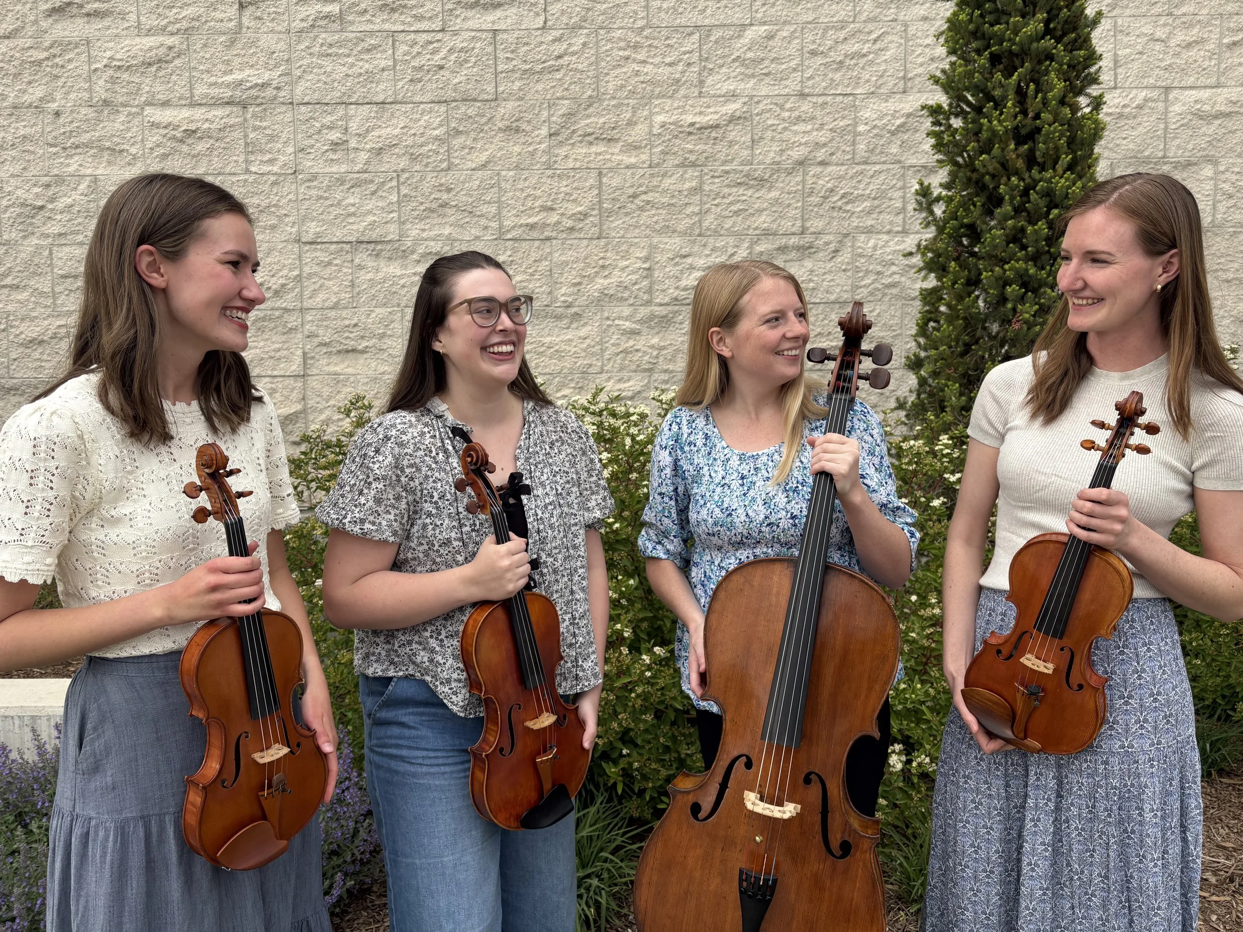 Five women standing outdoors, each holding a violin, and smiling at each other. They are dressed in casual clothing, with a textured beige brick wall and green bushes in the background.