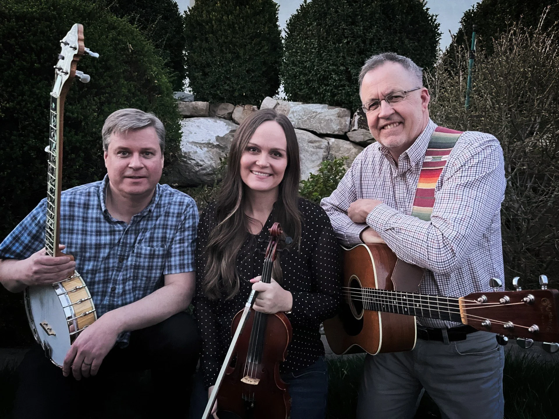 Three people, two men and one woman, posing outdoors with musical instruments. The man on the left is holding an electric guitar, the woman in the middle is holding a violin, and the man on the right is holding an acoustic guitar. They are standing in front of some bushes and rocks.