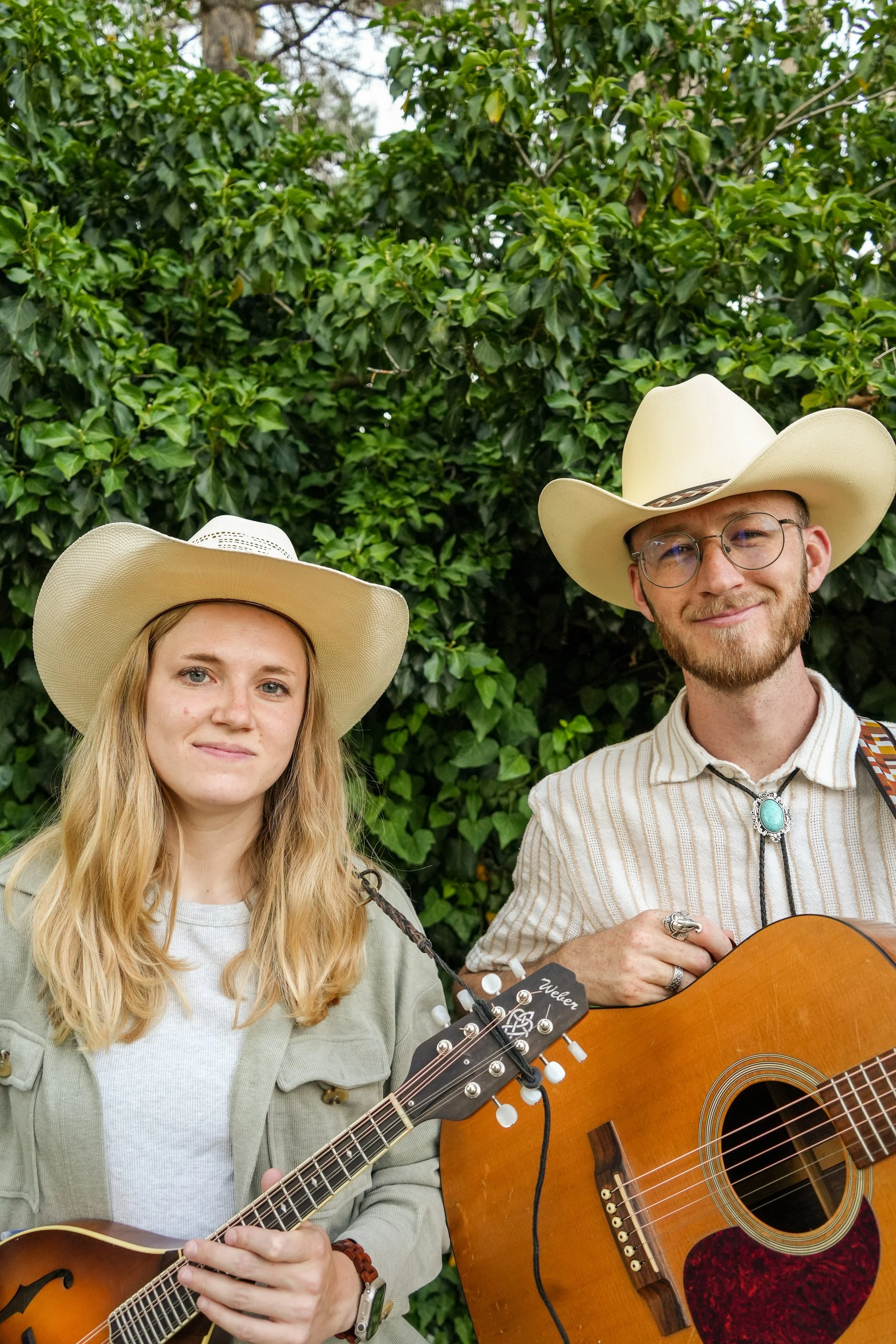 Two young musicians, a woman and a man, wearing cowboy hats, playing guitars outdoors in front of green foliage.