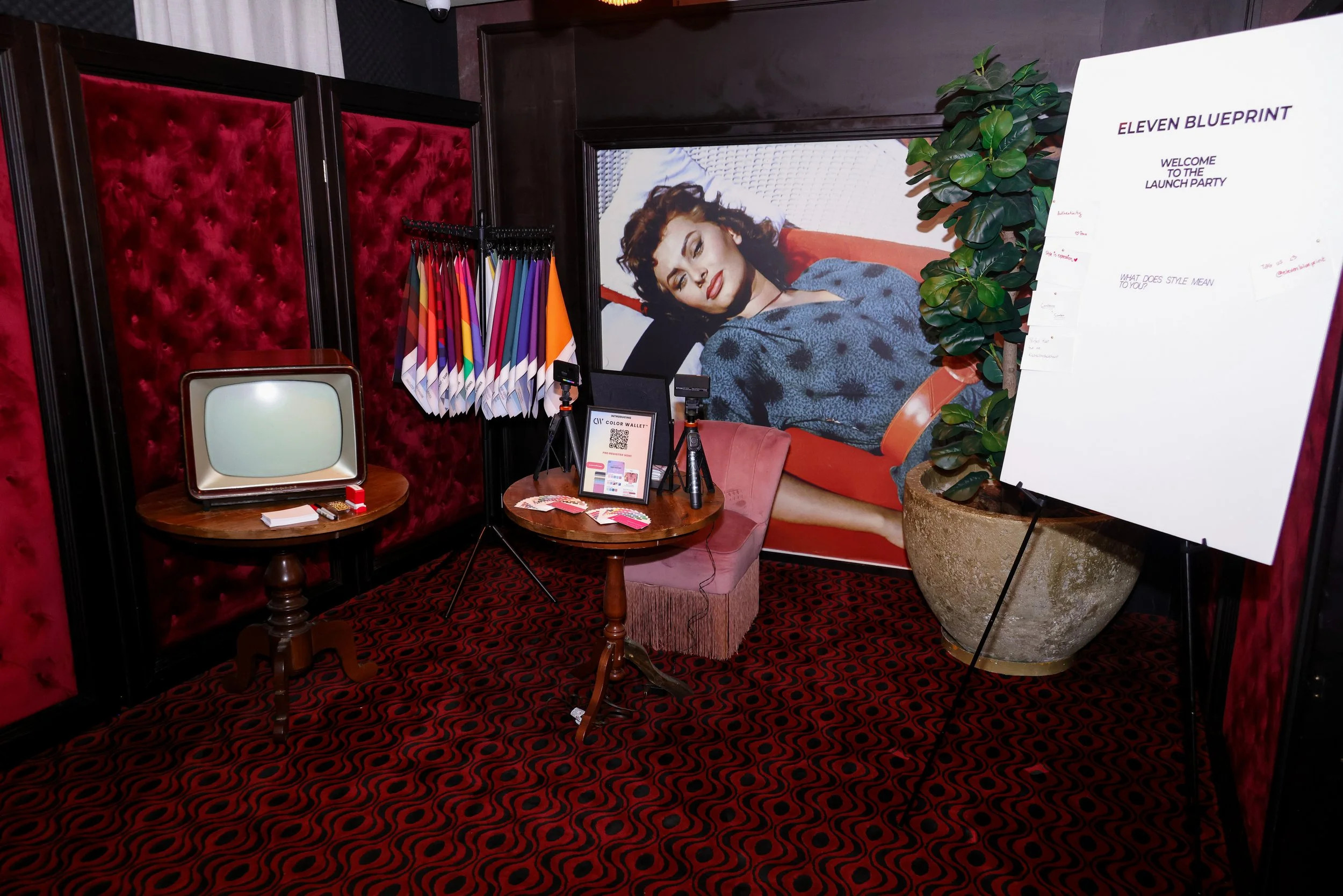 An interior scene of a room decorated with red velvet walls and patterned red and black carpet. There are colorful fabric swatches hanging on a rack, a small vintage television on a round wooden table, and a framed photo or screen showing a woman wit