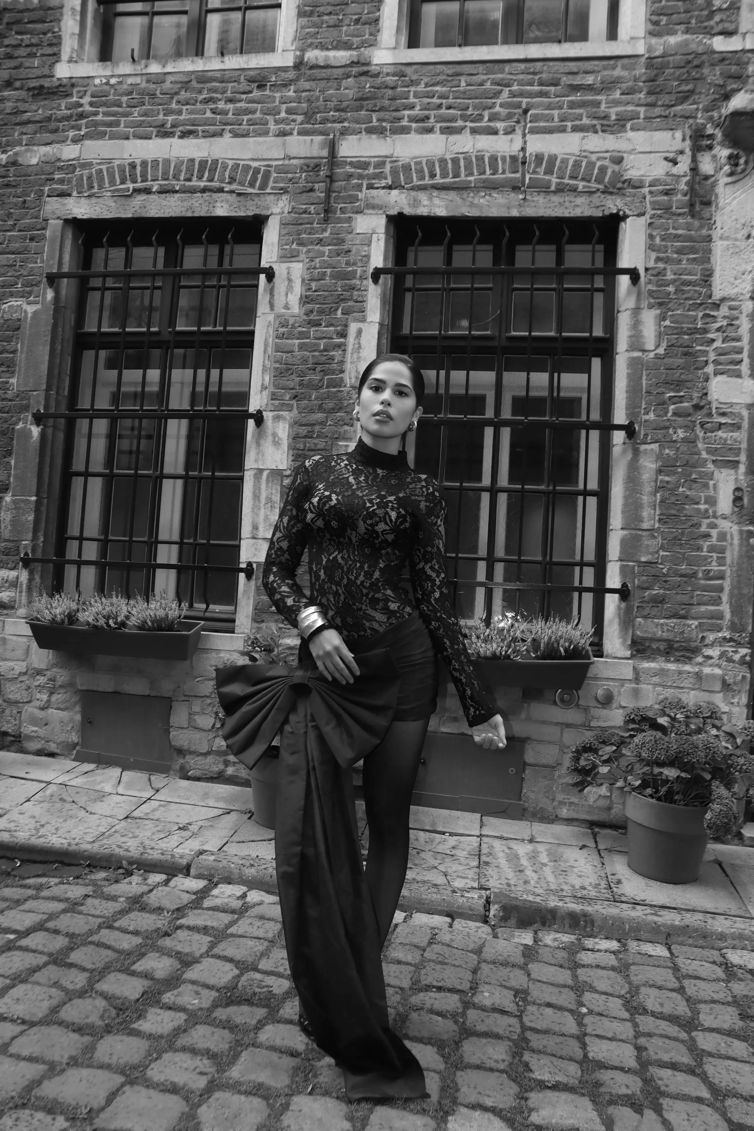 A woman in a black lace top, black shorts, and sitting on a bridge of big fabric with a bow stands on a cobblestone street in front of a brick building with barred windows and potted plants.