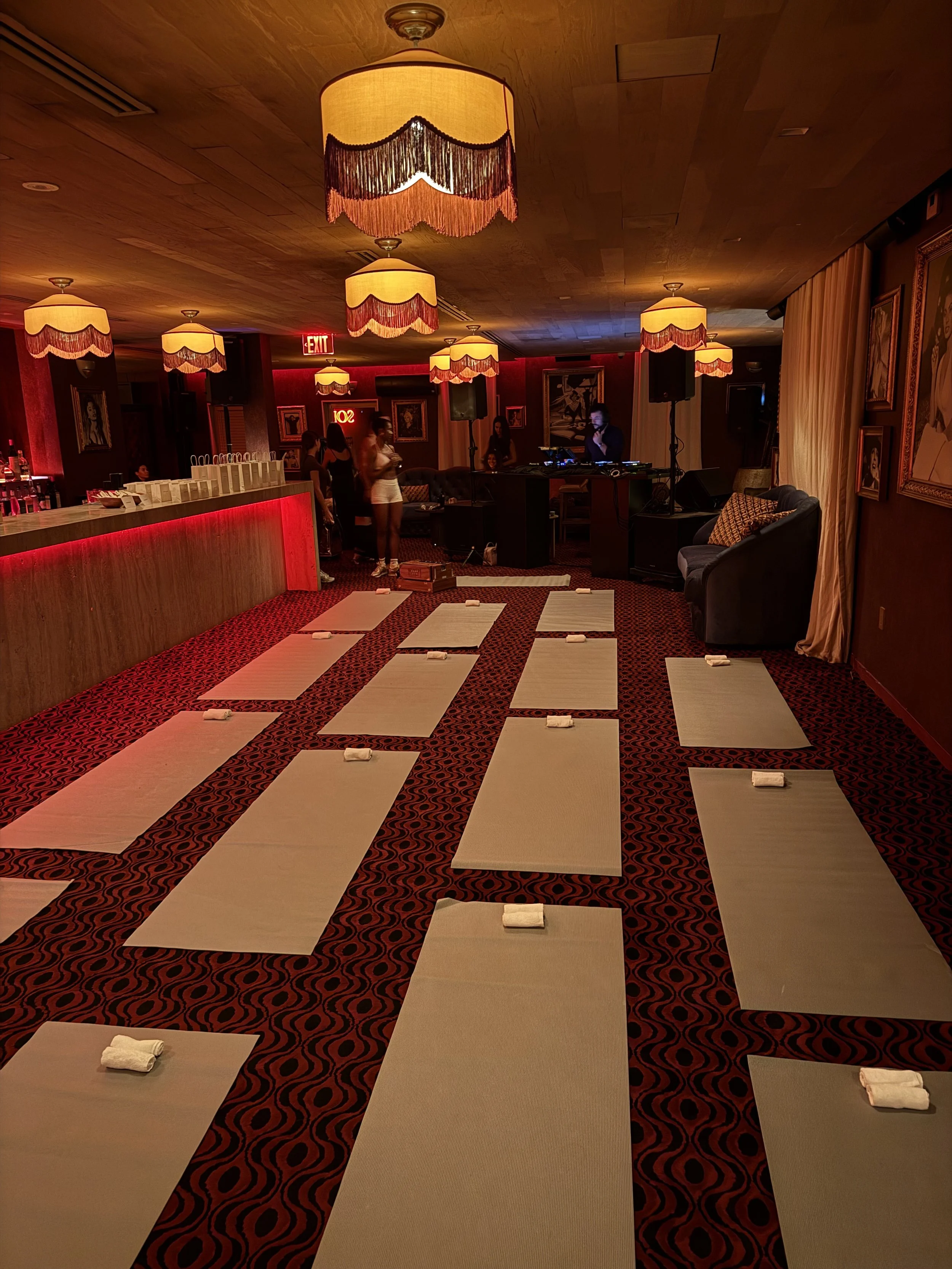 A dimly lit event space with yoga mats and small towels arranged on a red patterned carpet, decorative hanging lamps, a DJ setup, and a bar area with group of people gathered.