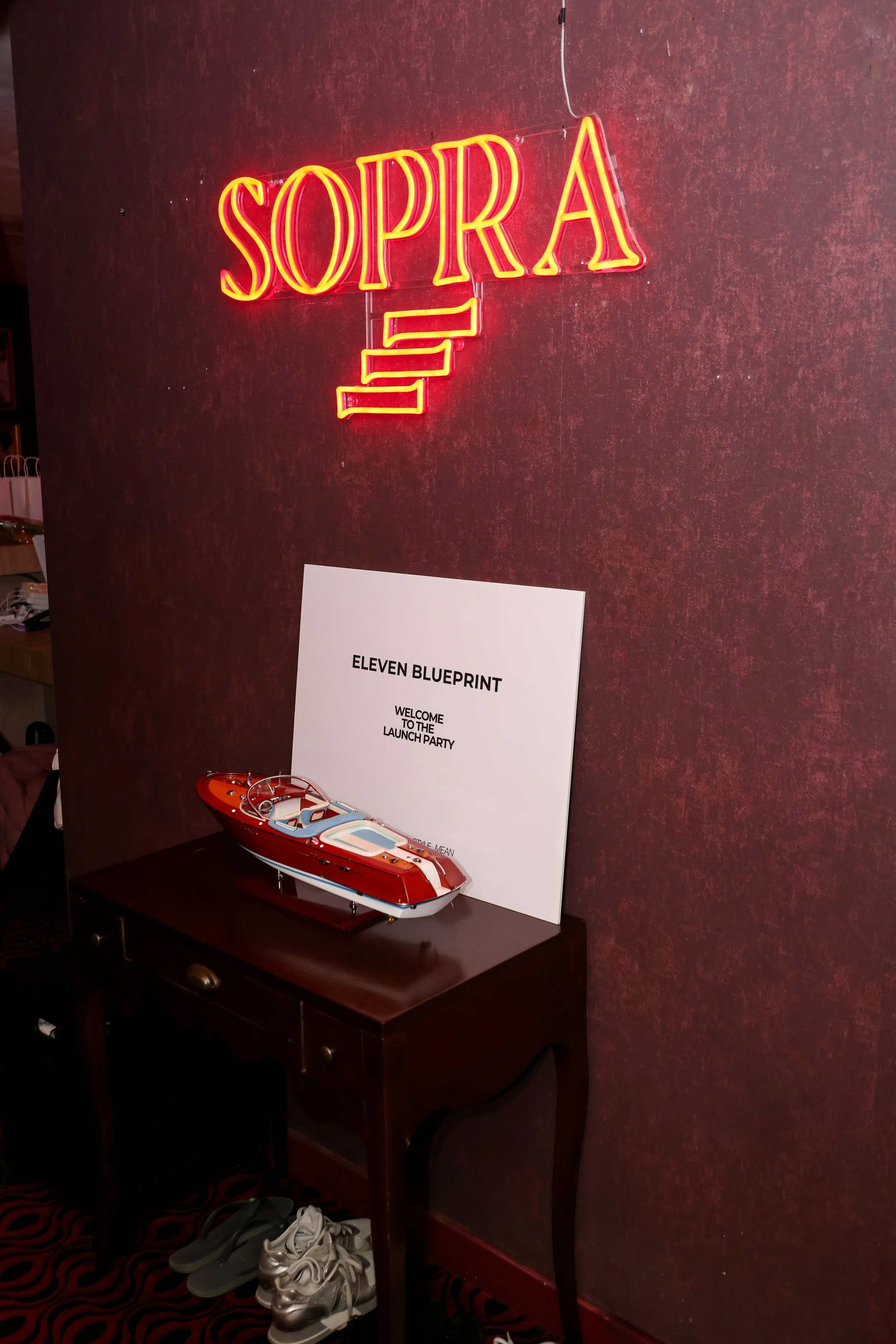 Neon sign spelling 'SOPRA' in red, a white sign saying 'ELEVEN BLUEPRINT', and a model boat on a dark wooden table against a textured red wall.