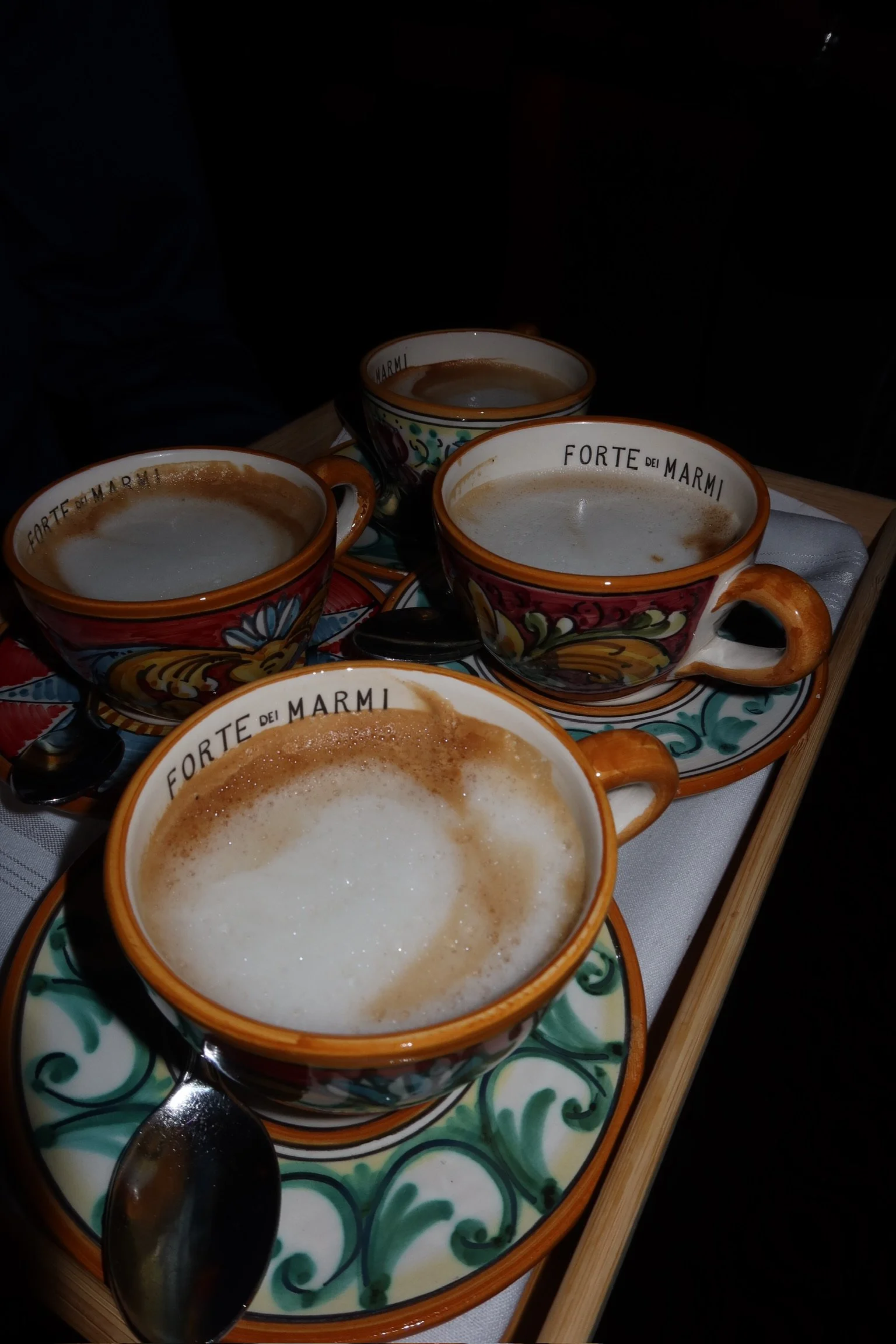 Four ceramic cups of Italian-style coffee with foam, placed on colorful saucers, on a wooden tray with spoons, with a white cloth underneath.