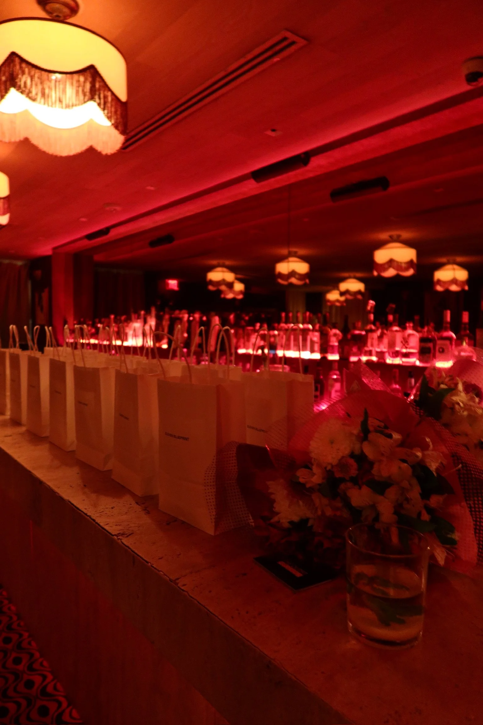 A row of gift bags and a flower bouquet on a bar or counter with bottles of alcohol illuminated by warm, red lighting in a nightclub or lounge setting.