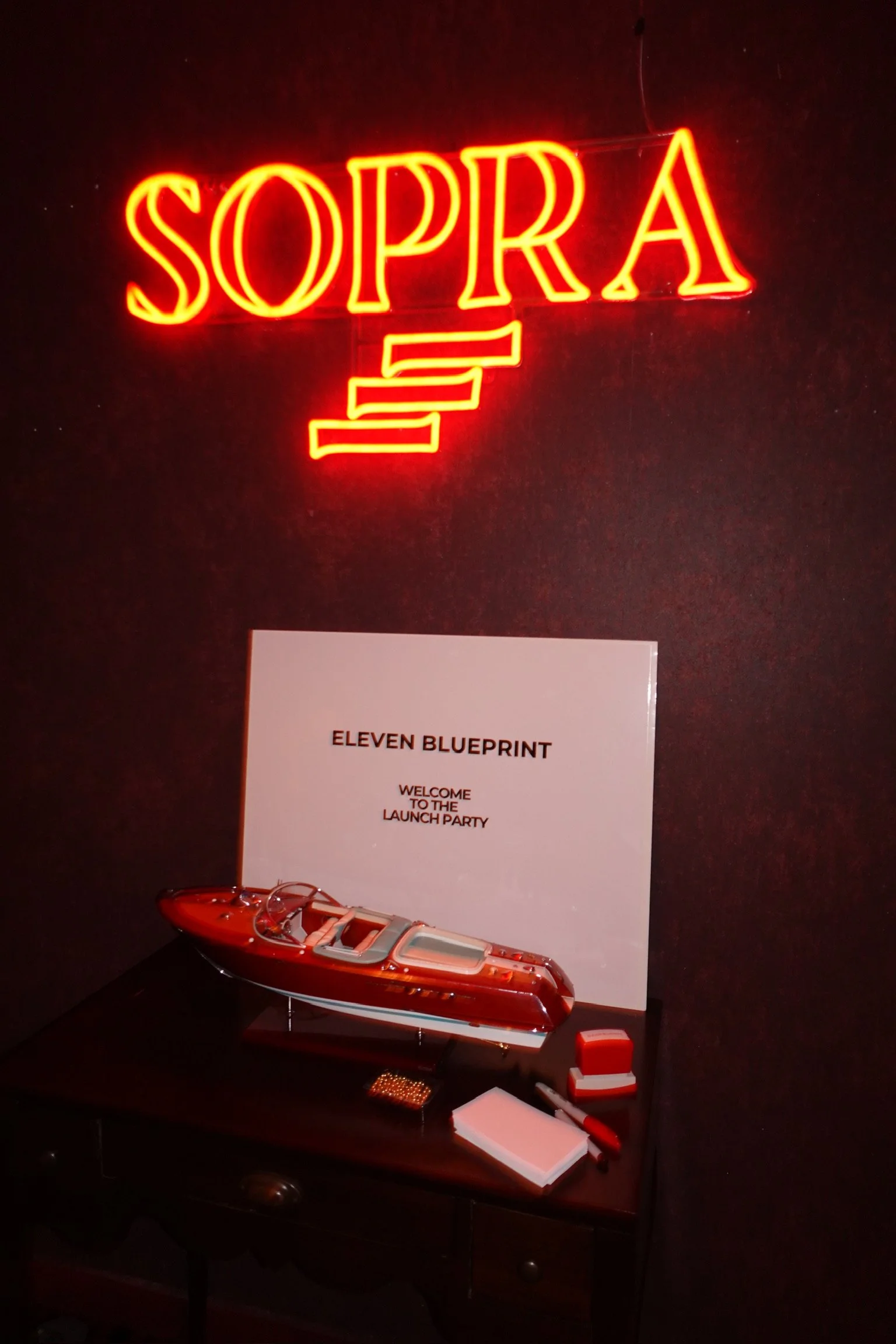 Red illuminated sign reading 'SOPRA' hangs on a dark wall. Below it, a table displays a model boat, a notepad, a pen, a small red box, and a sheet of paper with the title 'ELEVEN BLUEPRINT' and the subtitle 'WELCOME TO THE LAUNCH PARTY'.