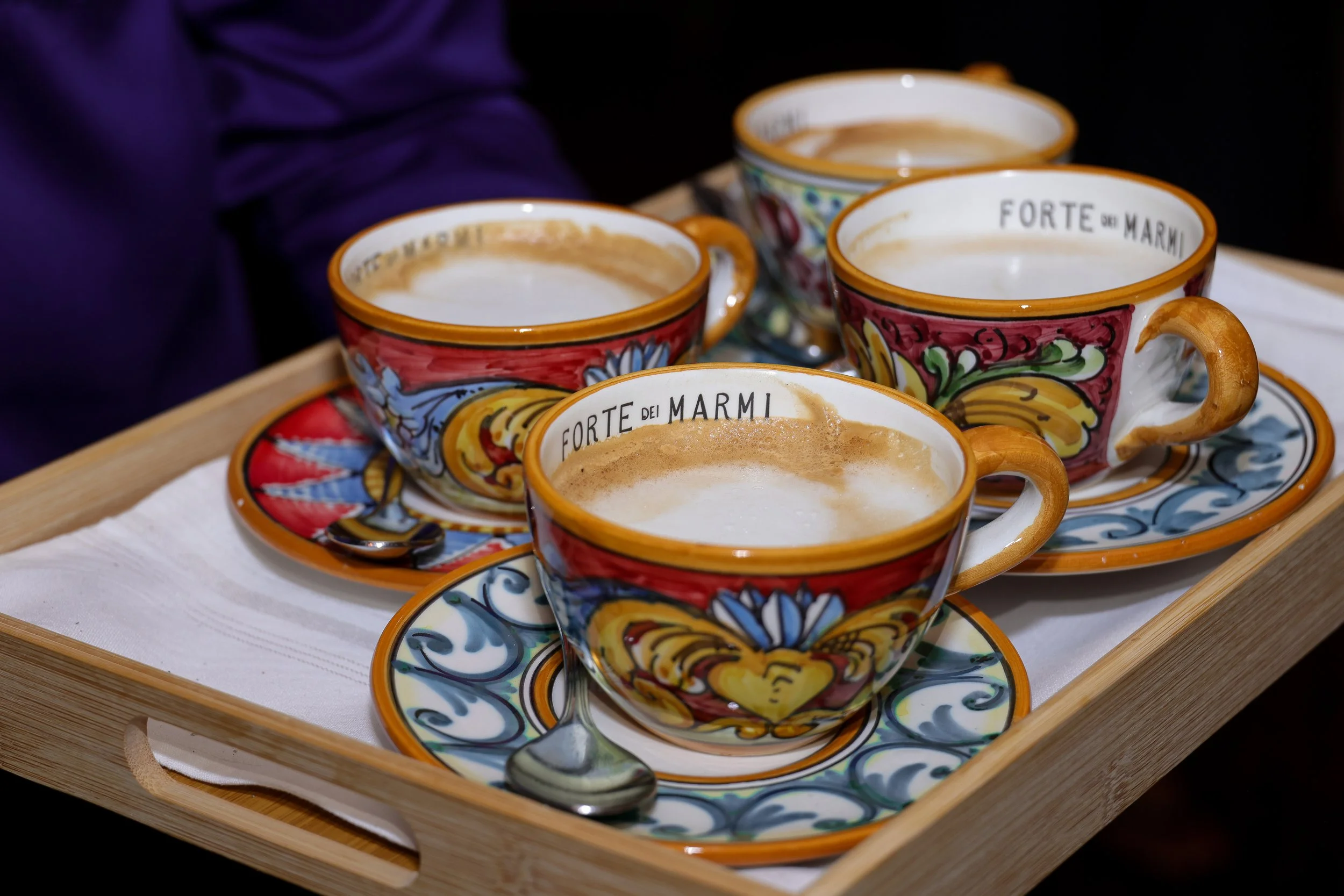 Four colorful ceramic cups of coffee on a wooden tray, each on a matching saucer, with spoons inside, with a purple sleeve in the background.