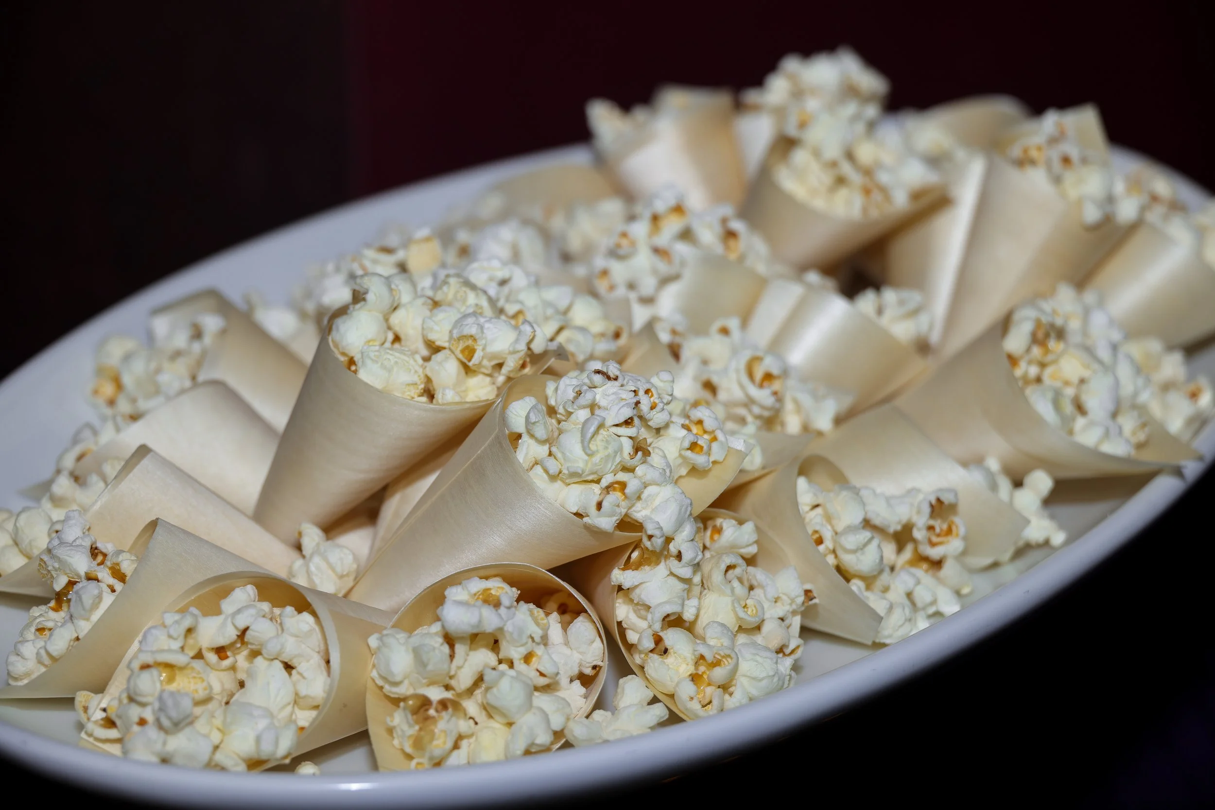 A white oval bowl filled with paper cones of popcorn. The popcorn is freshly popped and white, with some golden kernels, arranged closely together.