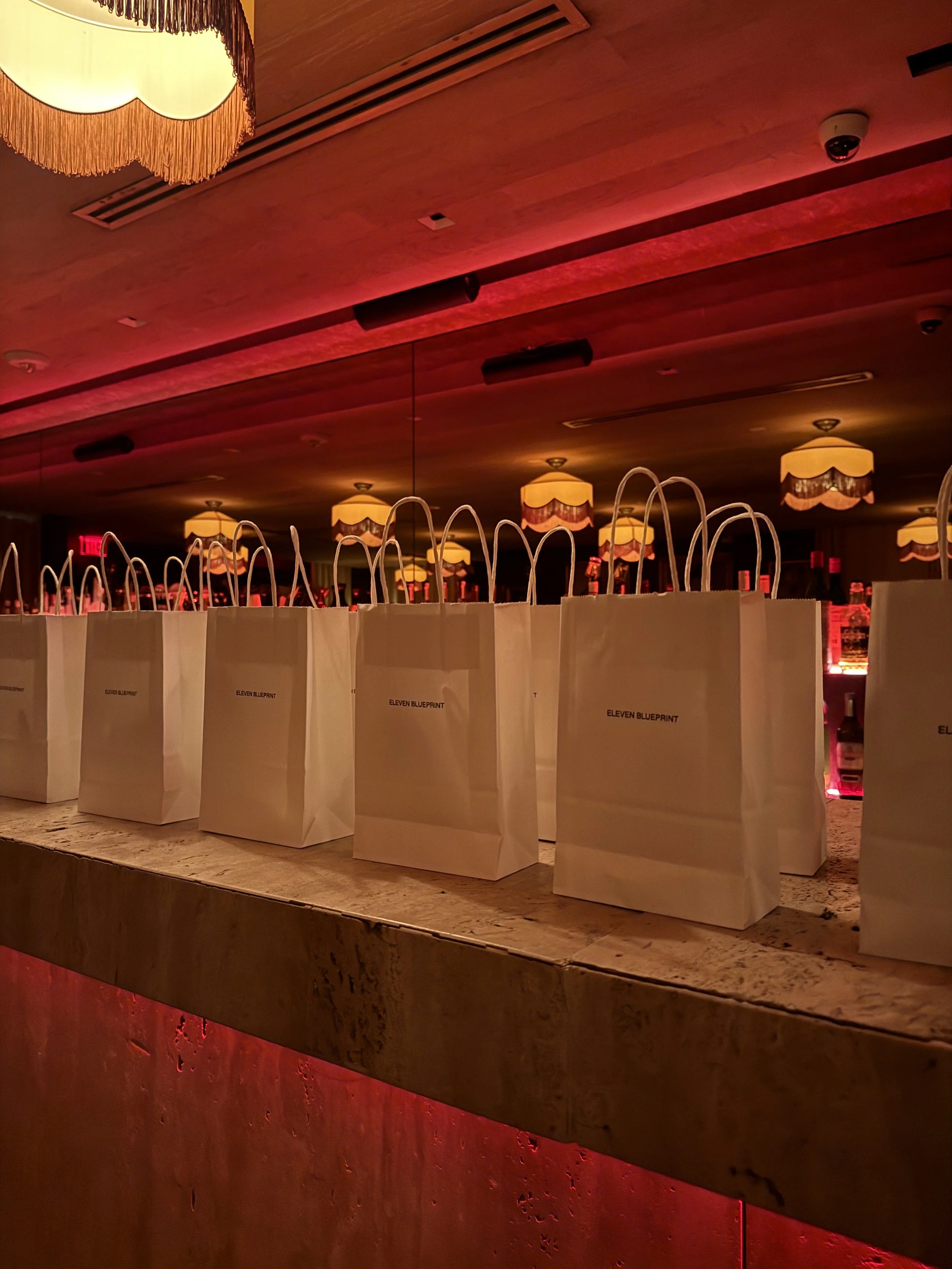 Several white paper bags labeled 'Eleven Blueprint' arranged on a bar counter in a dimly lit room with decorative hanging lamps and red ambient lighting.
