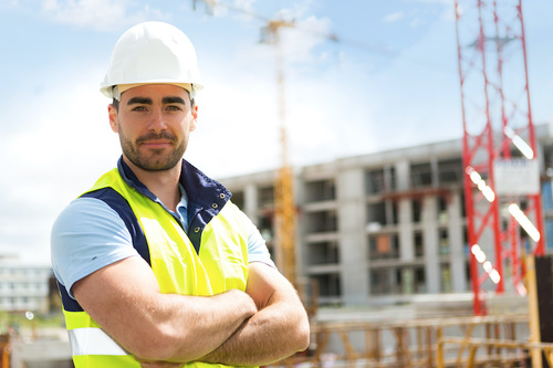 A male construction worker wearing a white hard hat and a yellow safety vest stands with arms crossed at a construction site during daytime.