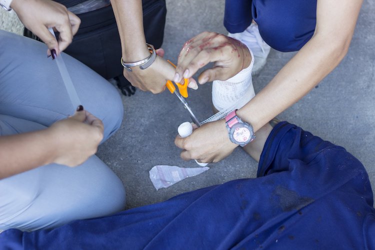 Medical personnel assist a person with a foot injury, using scissors and a bandage, while others help stabilize the foot.