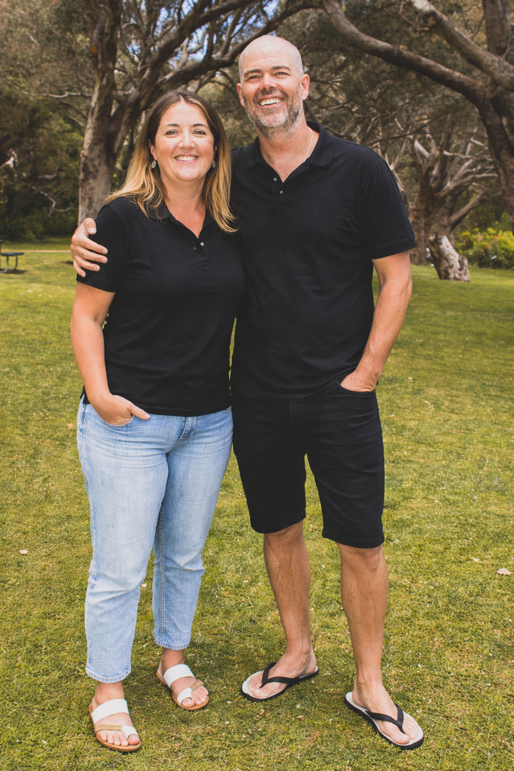 A smiling couple with their arms around each other, standing on a grassy park with large trees in the background.