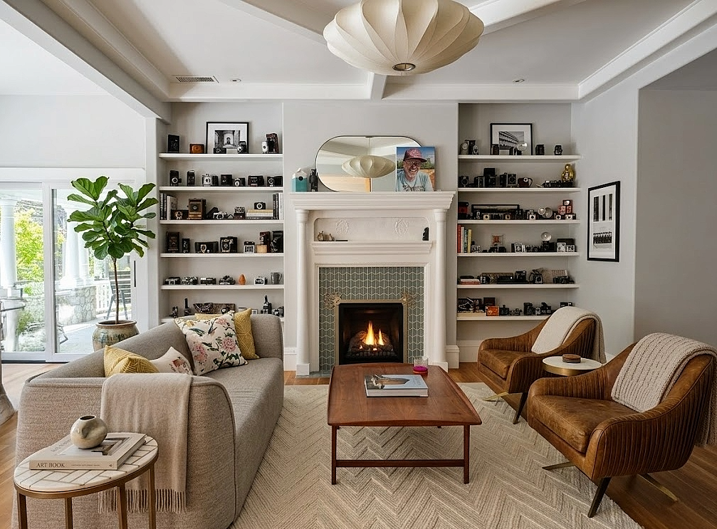 Living room with a fireplace, white walls, built-in shelves with cameras and decor, a large window/door with a view of greenery, a gray sofa with colorful pillows, two brown armchairs, a wooden coffee table, a mirror above the fireplace, and leaning photos on the walls.