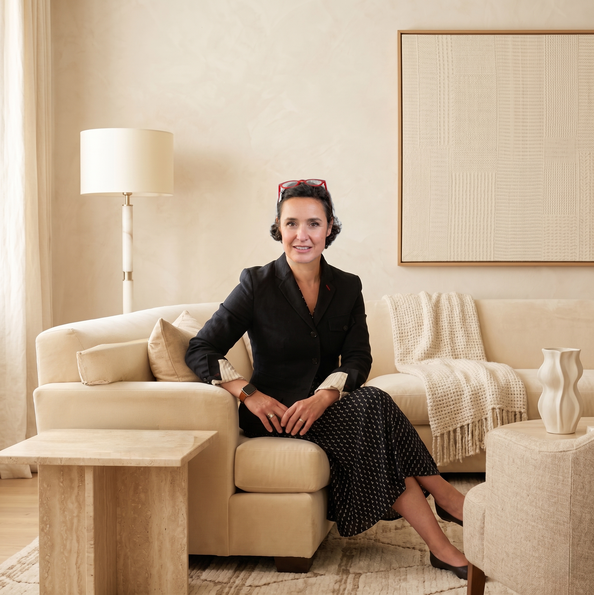 A woman with short dark curly hair and red glasses on her head, wearing a black jacket, sitting on a beige sofa in a room with light-colored walls, a white floor lamp, and a wooden framed decorative piece on the wall.