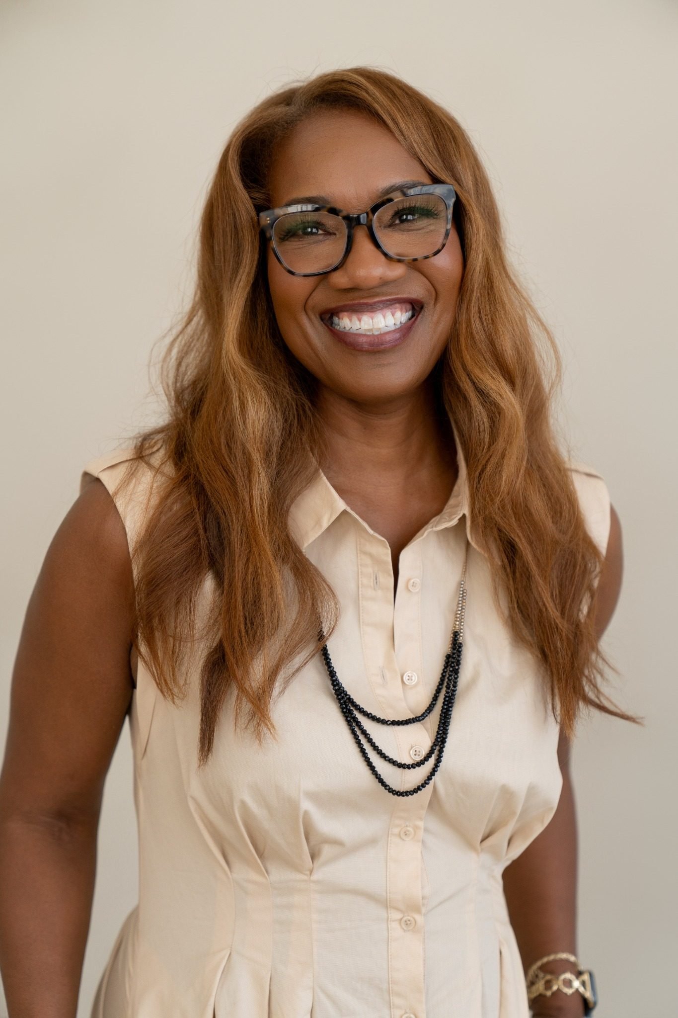 A smiling woman with brown hair, wearing glasses, a sleeveless beige buttoned dress, layered beaded necklaces, and a gold bracelet, stands against a plain background.
