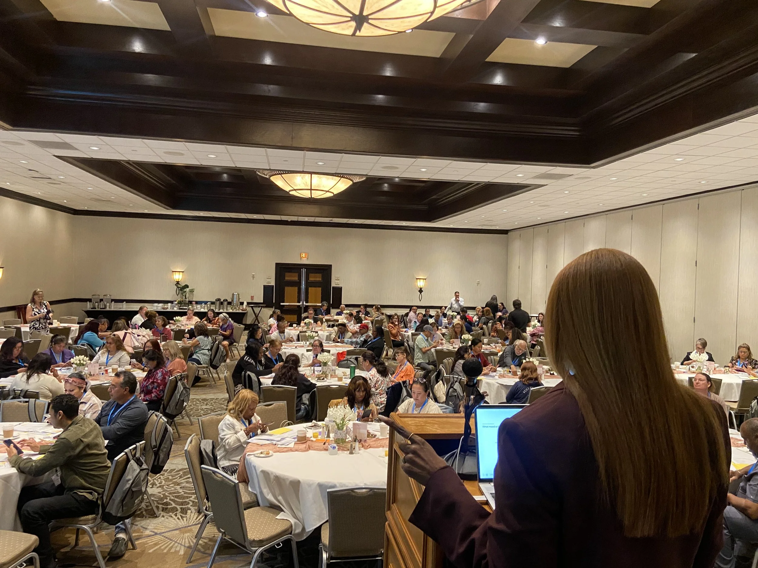 A large conference room filled with people sitting at round tables, listening to a speaker at a podium. The room has a coffered ceiling with large ornate light fixtures and wall-mounted lights. Attendees are using laptops, phones, and note-taking materials.
