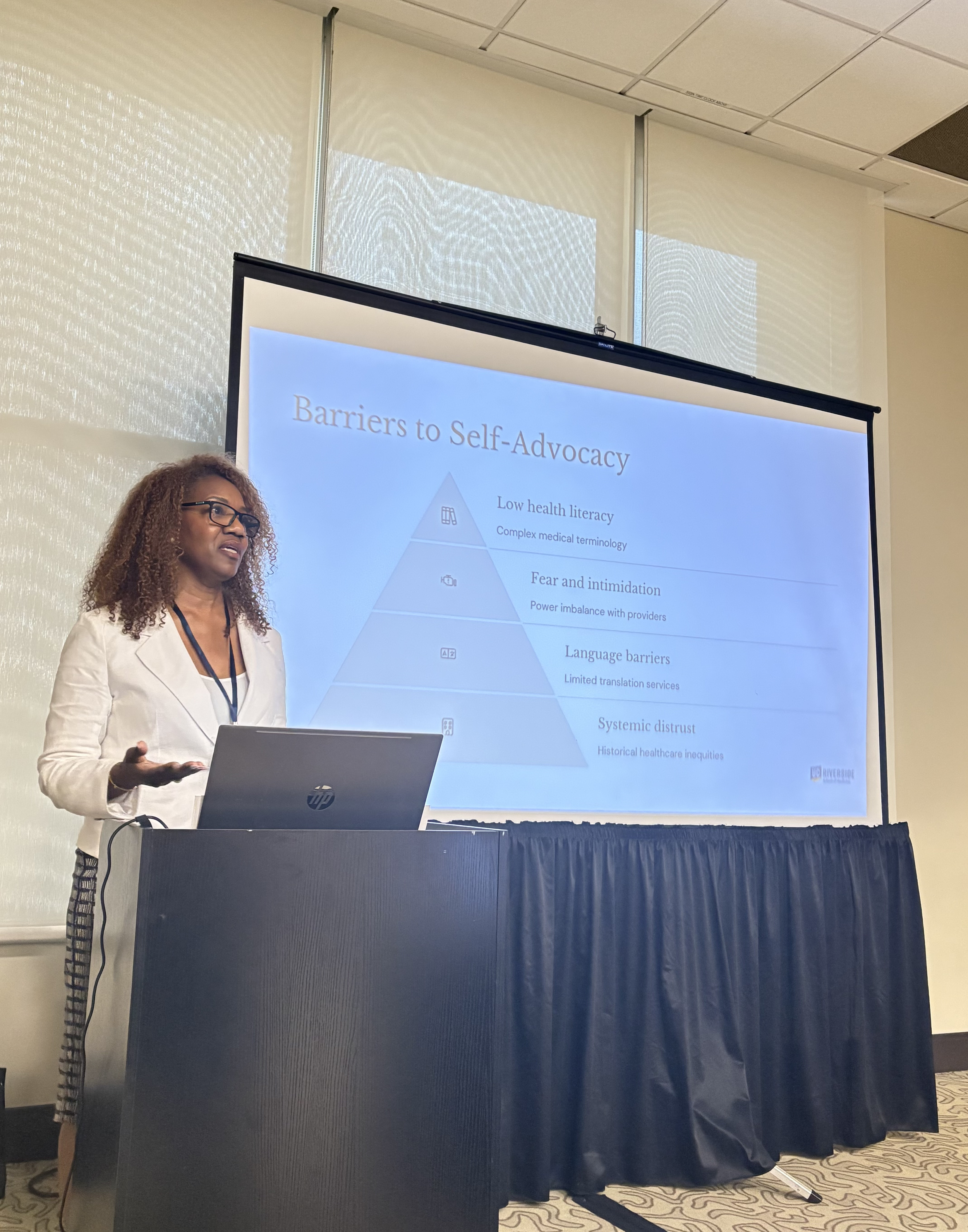 Dr. Michelle Christine, with glasses speaks at a podium beside a projected slide titled “Barriers to Self-Advocacy,” in a bright conference room; the mood is educational.