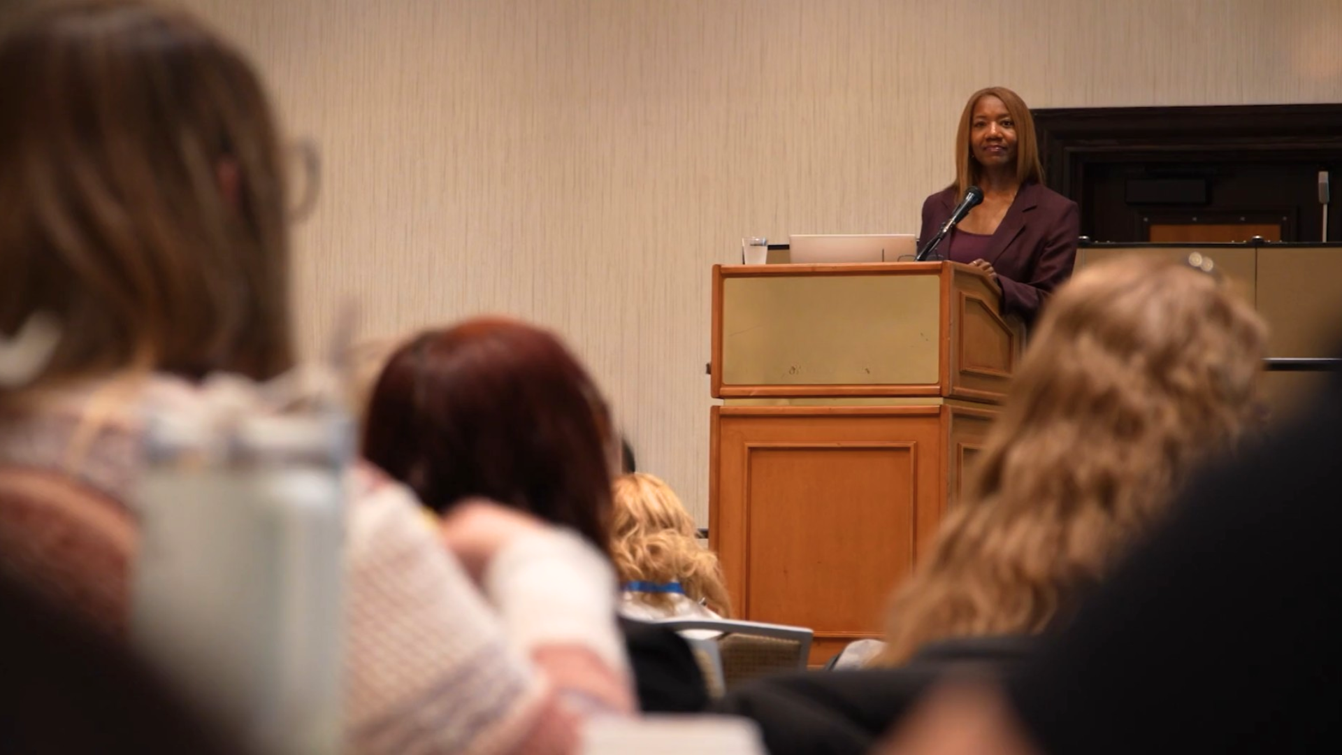 A smiling Dr. Michelle Christine speaks at a wooden podium at caregiver training summit, addressing a seated audience in a focused, professional setting.