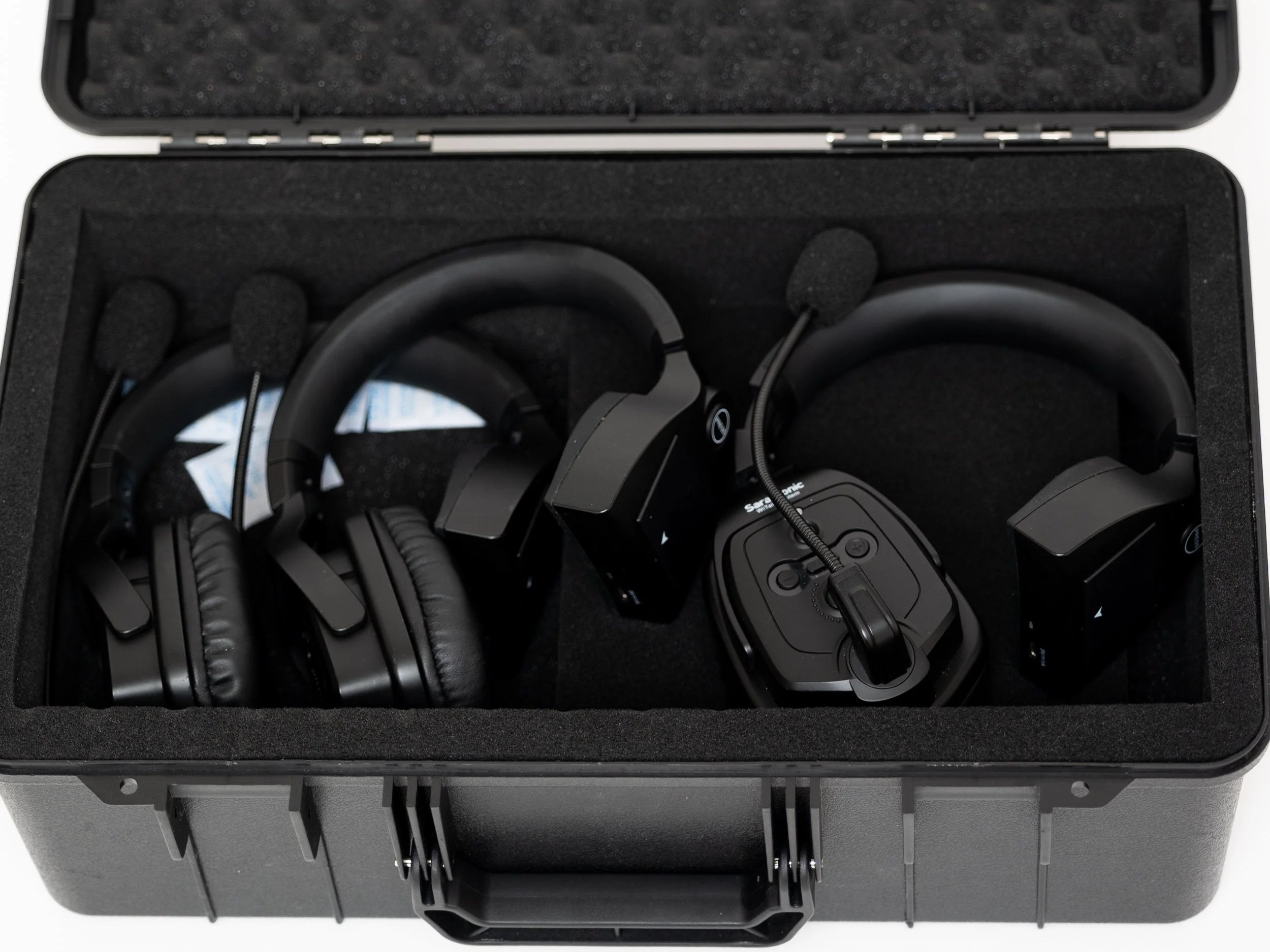 A black carrying case containing three headsets with microphones. Two are larger with over-ear design, and one is smaller with a built-in control panel and microphone boom.
