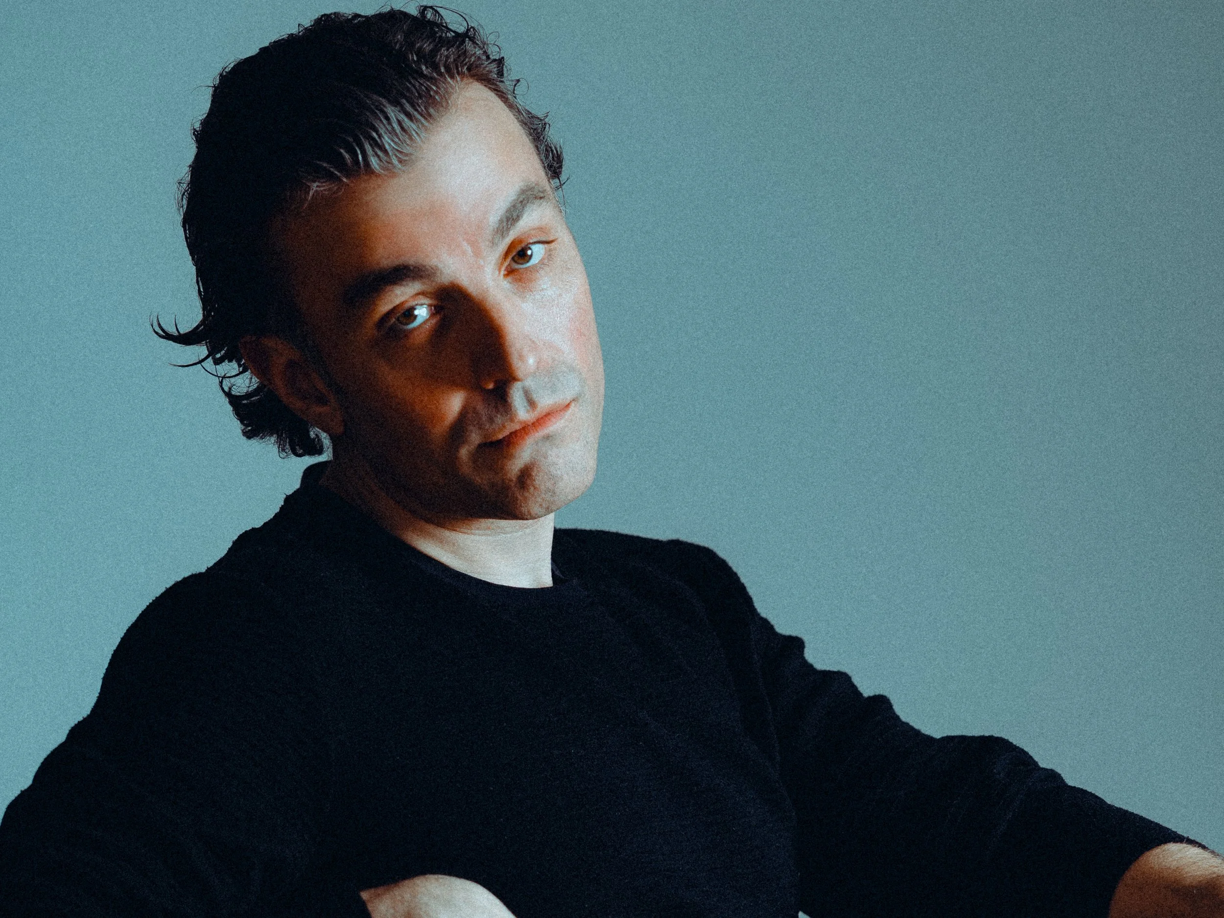 A man with dark hair, wearing a black shirt, looking at the camera with a neutral expression against a plain background.