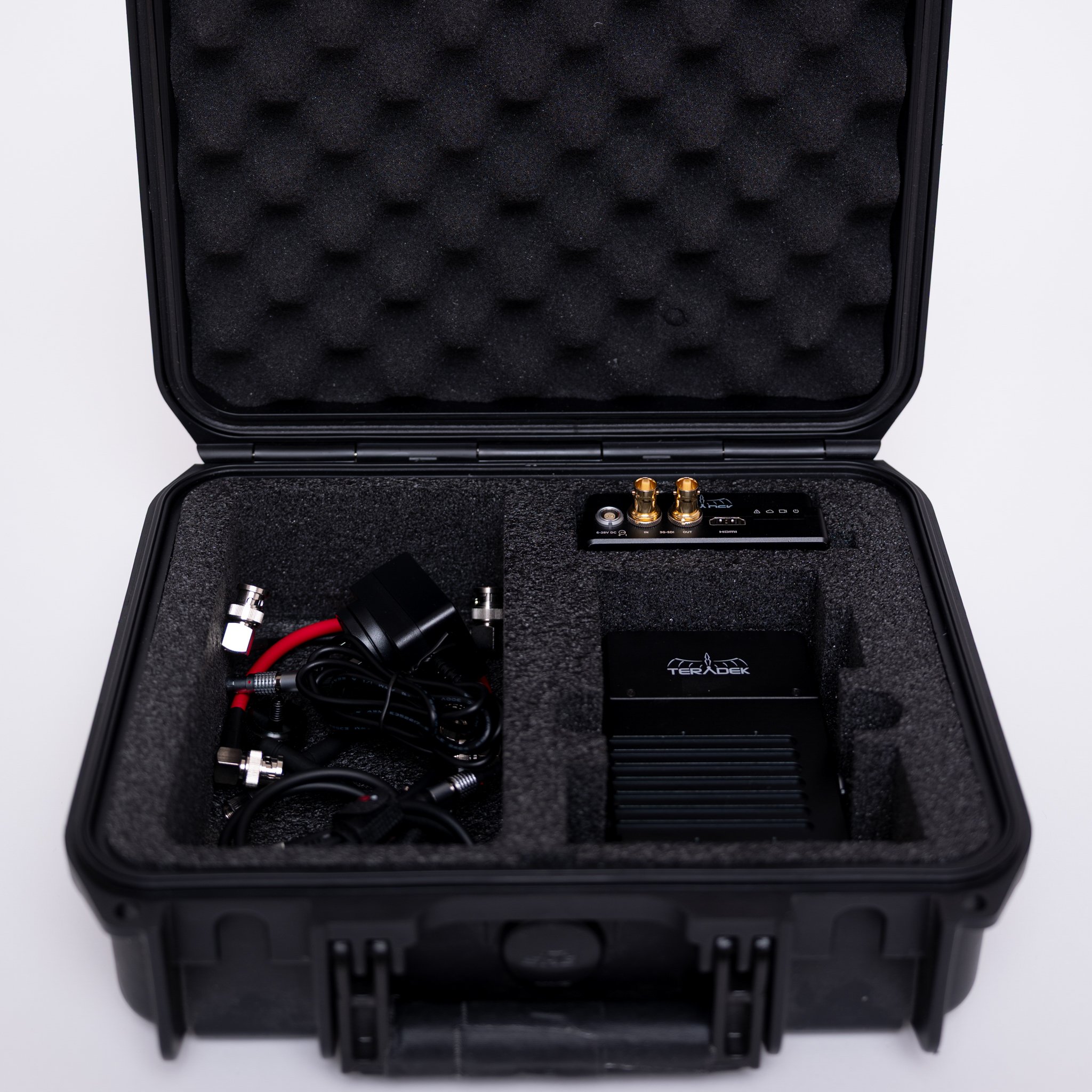 Open black protective case containing electronic equipment with foam padding, including wires, connectors, a small device with gold ports, and a black unit with the logo Teradek.
