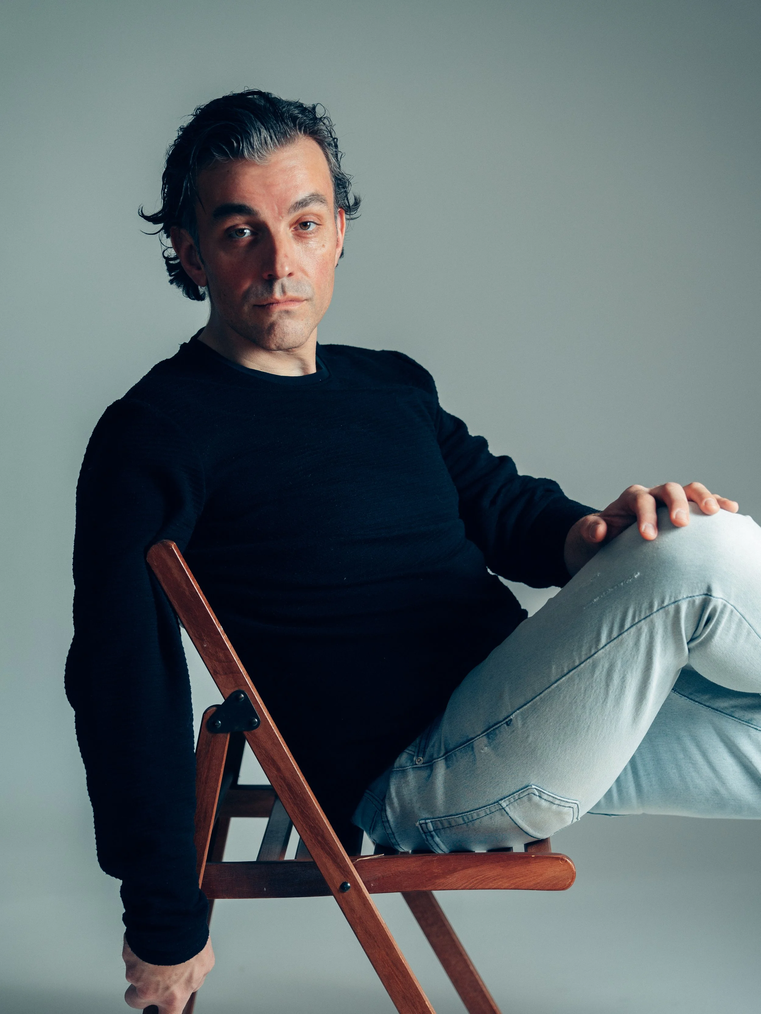 A man with dark hair and fair skin sitting on a wooden chair against a plain background, wearing a black shirt and light-colored jeans, looking directly at the camera.