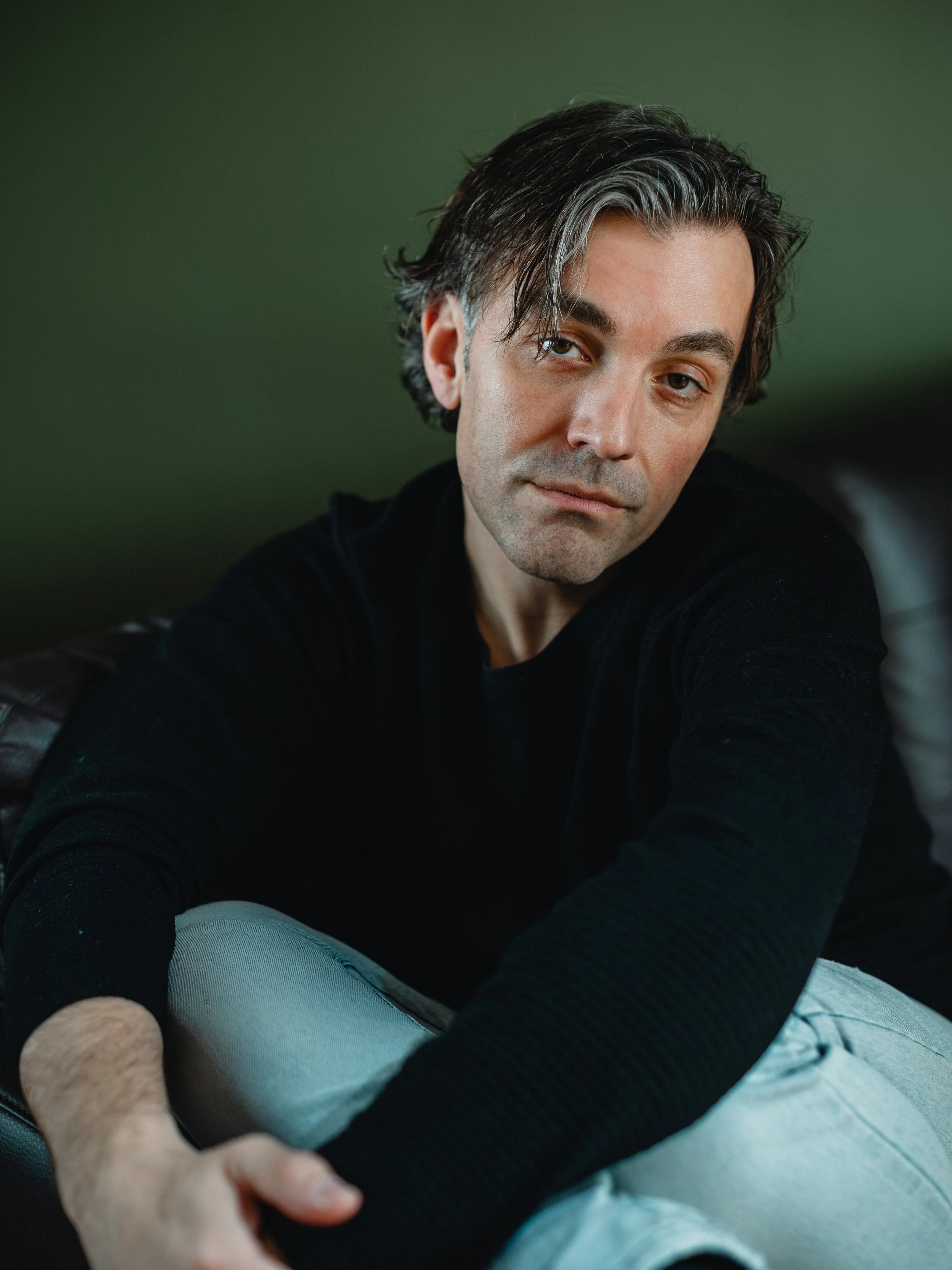 A man with dark wavy hair, wearing a black sweater, sitting on a black leather chair against a green background.