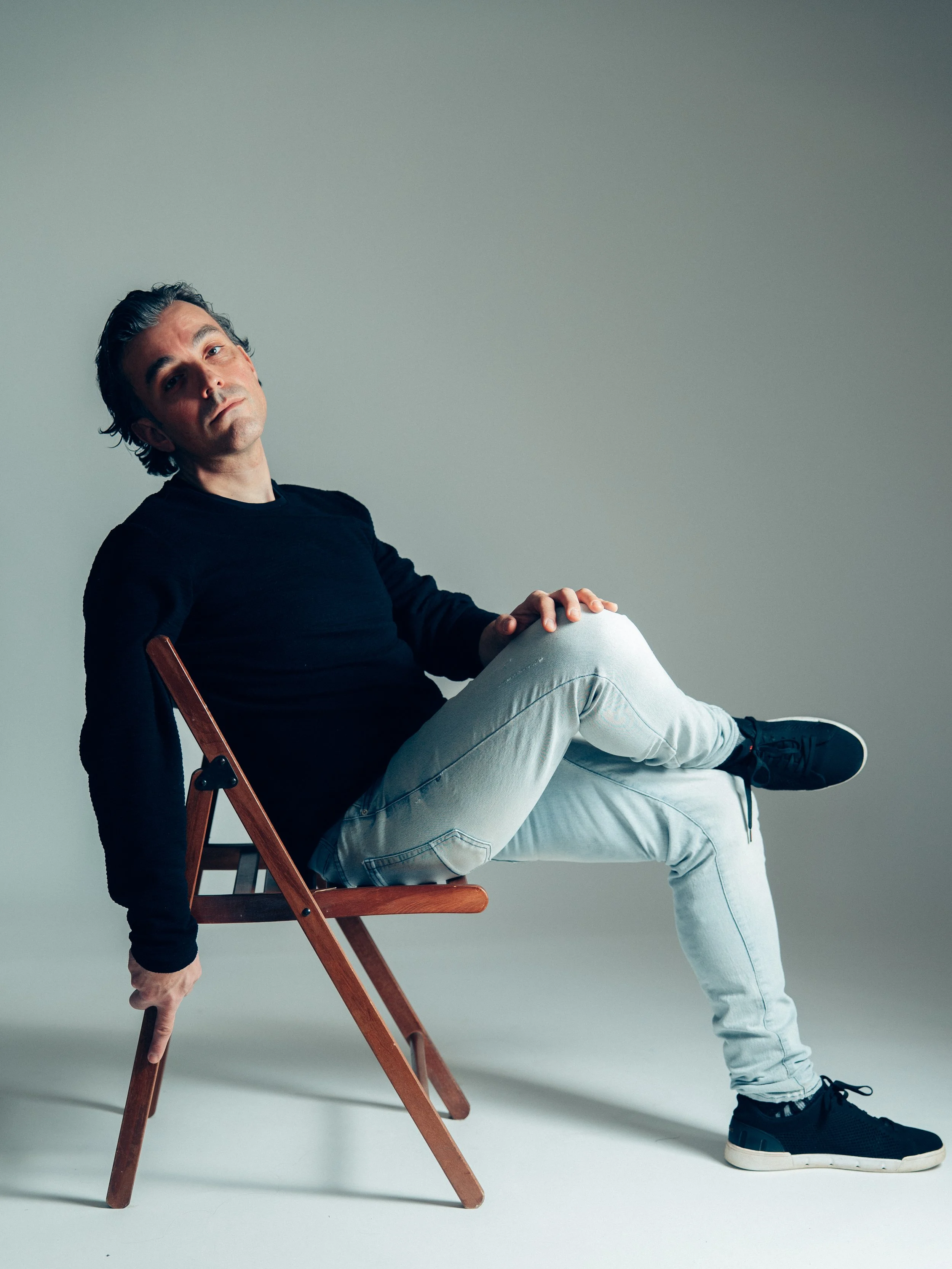 A man sitting sideways on a wooden chair with his legs extended and crossed. He is wearing a black shirt, light blue jeans, and black sneakers. The background is plain and gray.