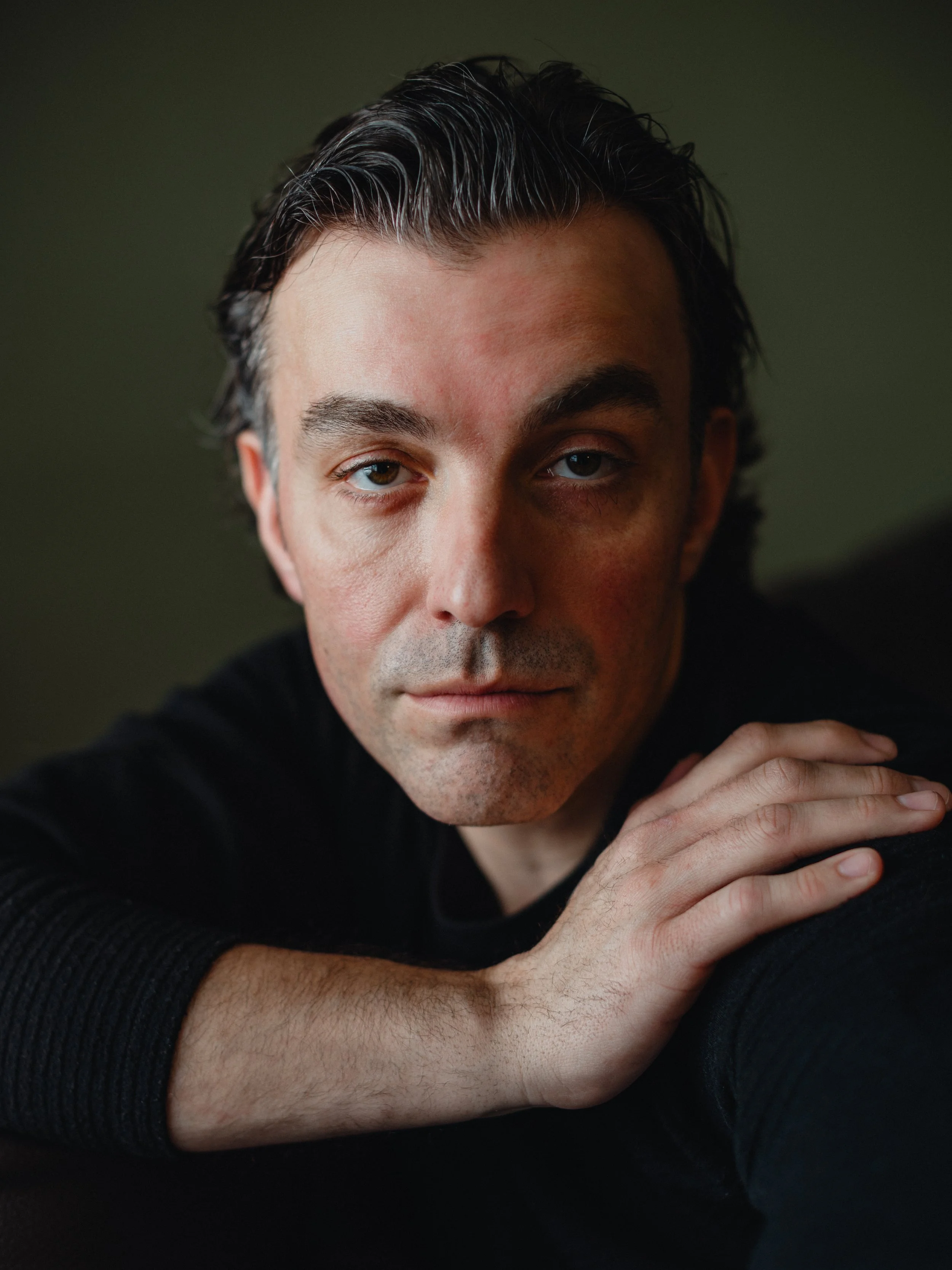Close-up portrait of a man with dark hair and blue eyes, wearing a black shirt, resting his head on his hand against a dark green background.