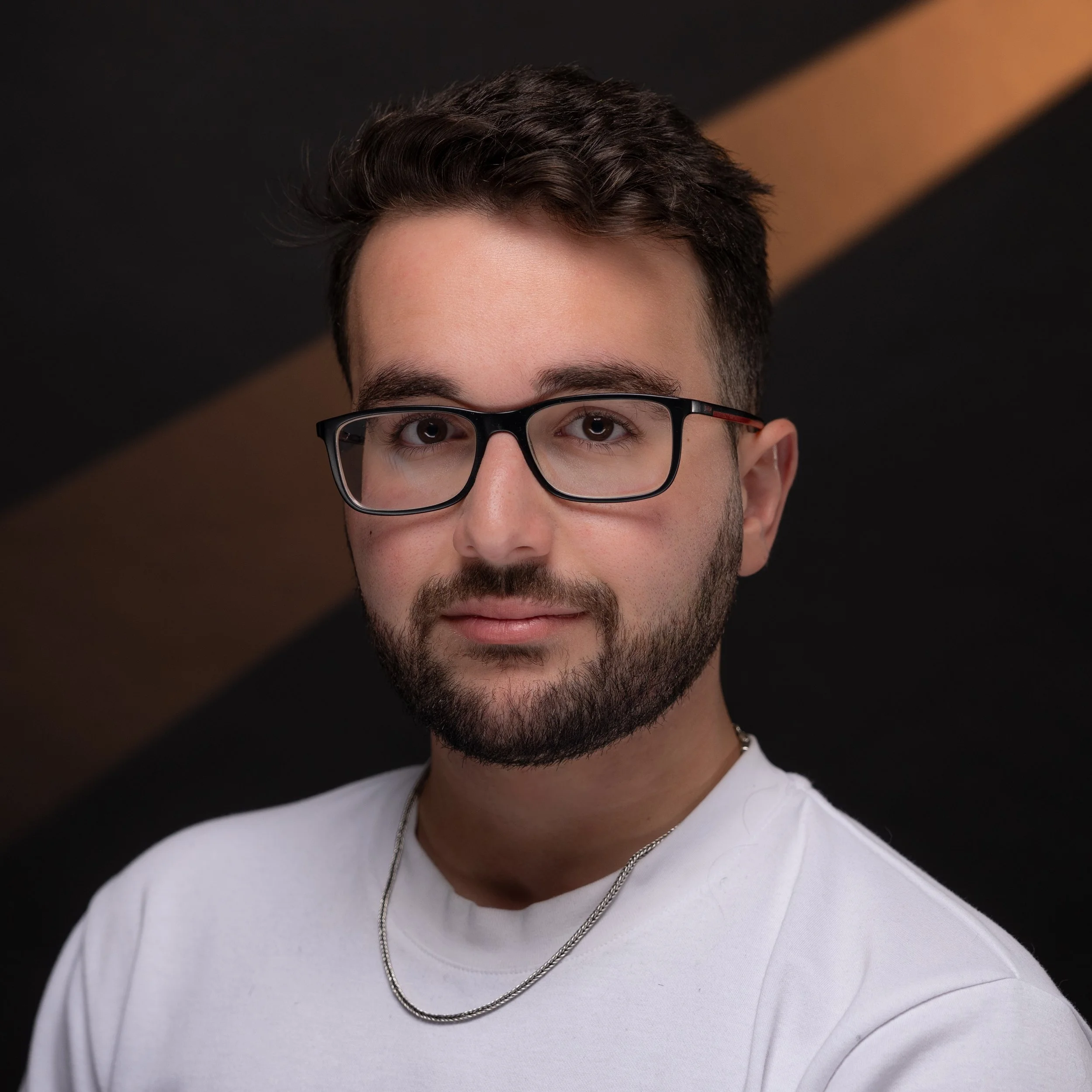 Portrait of a young man with glasses, short dark hair, beard, wearing a white shirt and a silver chain, smiling softly, against a black and brown background.