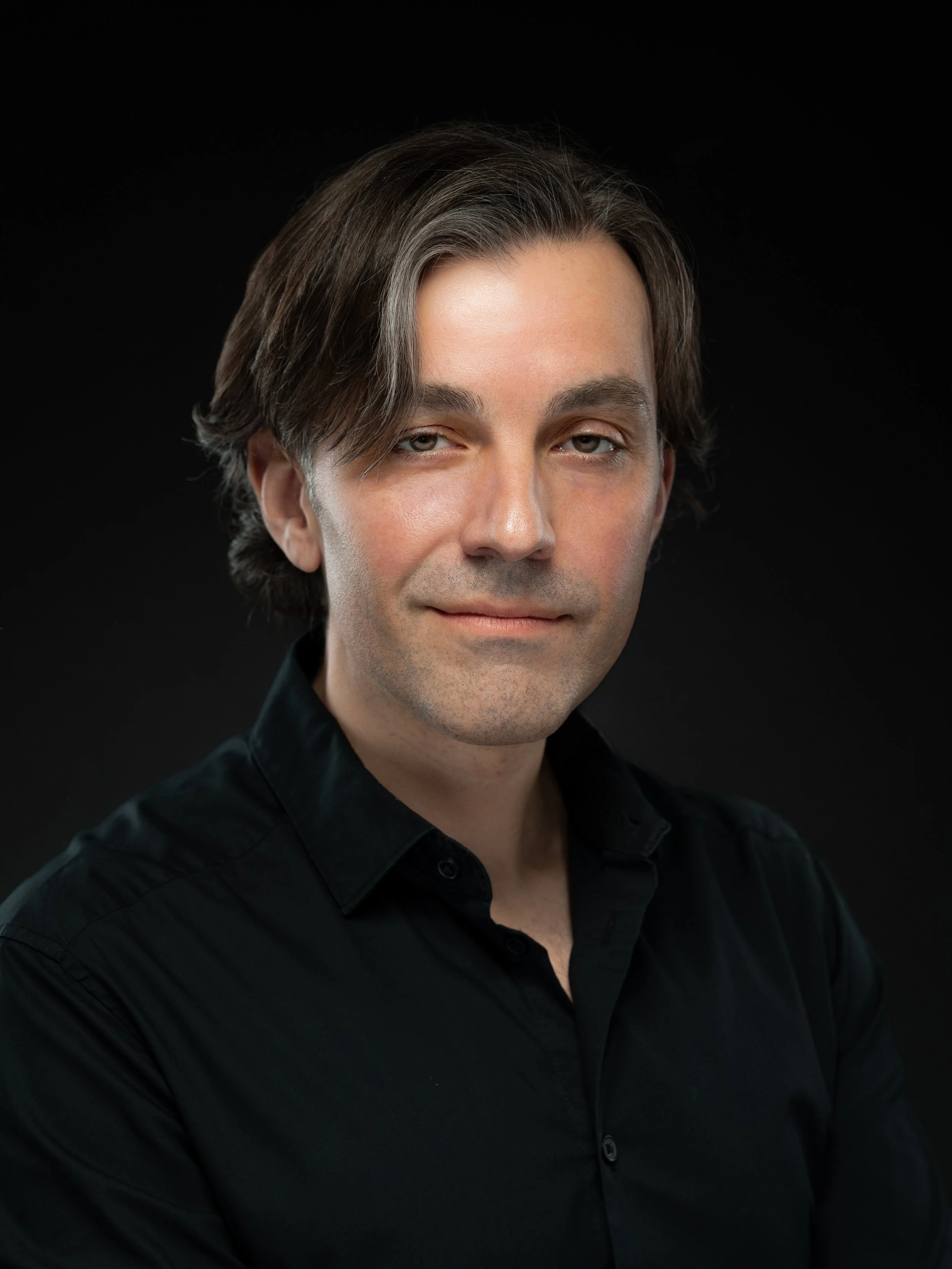 Portrait of a man with medium-length dark hair, wearing a black shirt, against a black background.