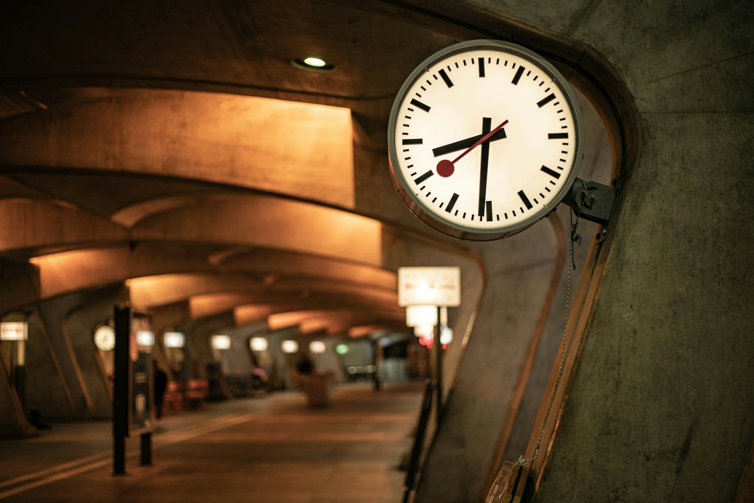 A large round clock shows the time as 7:07, hanging on a wall in an underground transit station with a curved ceiling and warm lighting.