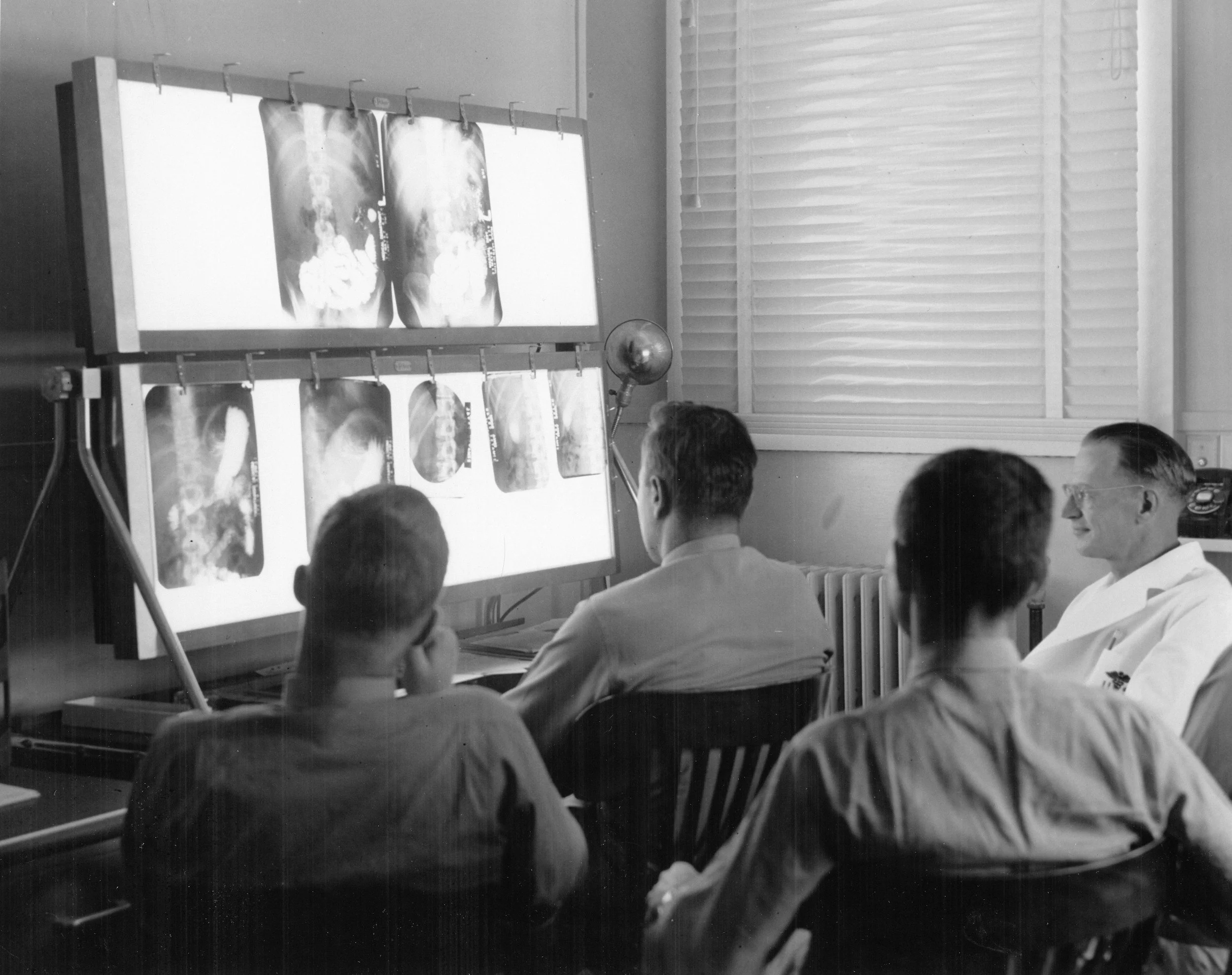 Four men, likely doctors, in a room with X-ray images displayed on a lightbox and a monitor, reviewing medical images together.
