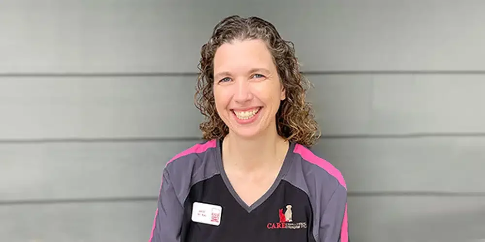 A woman with curly brown hair smiling standing against a gray wall, wearing a black and gray uniform with pink accents and a nametag.