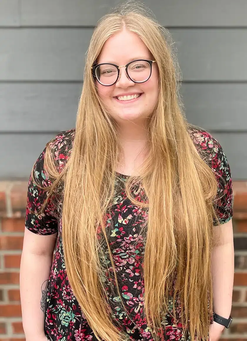 A young woman with long red hair, glasses, and a floral dress standing in front of a gray wall and a brick ledge, smiling at the camera.