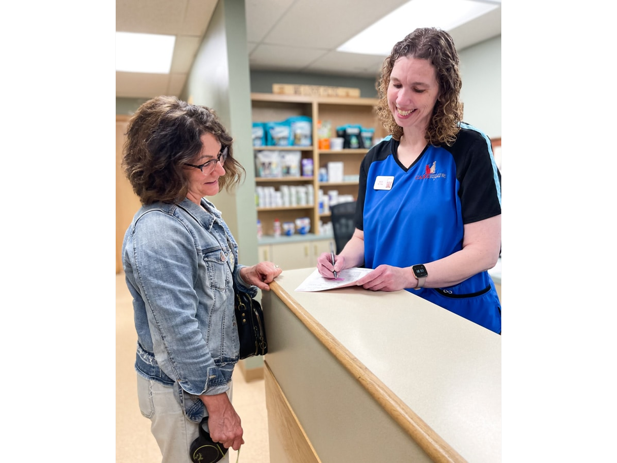 A woman in a denim jacket and beige pants checks in at a reception desk while interacting with a smiling female staff member in a blue uniform, holding a pen and writing on a form. The background includes shelves with medications and supplies.