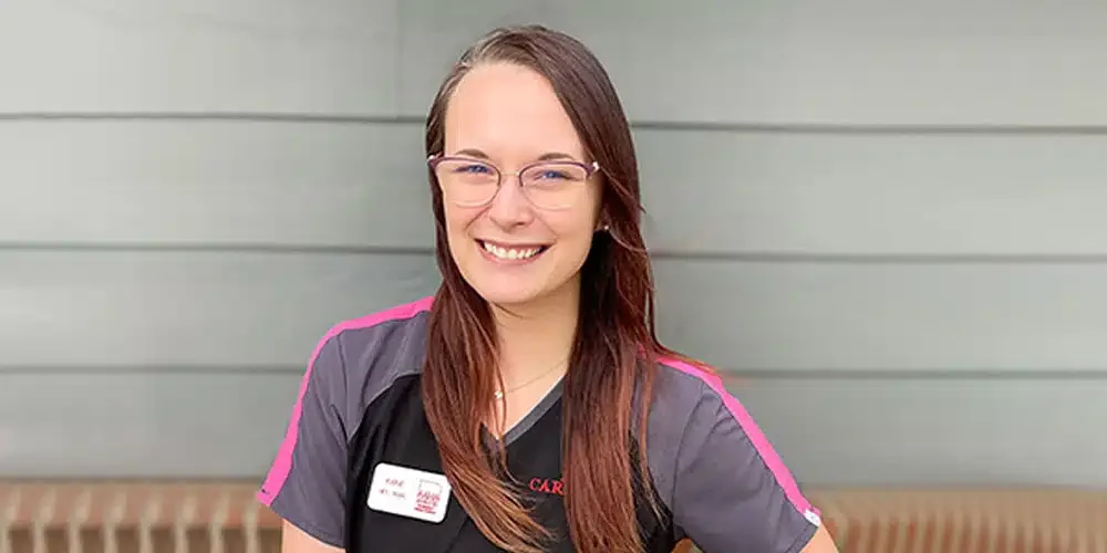Young woman with long brown hair, glasses, and a smile, standing outdoors in front of a gray wall.