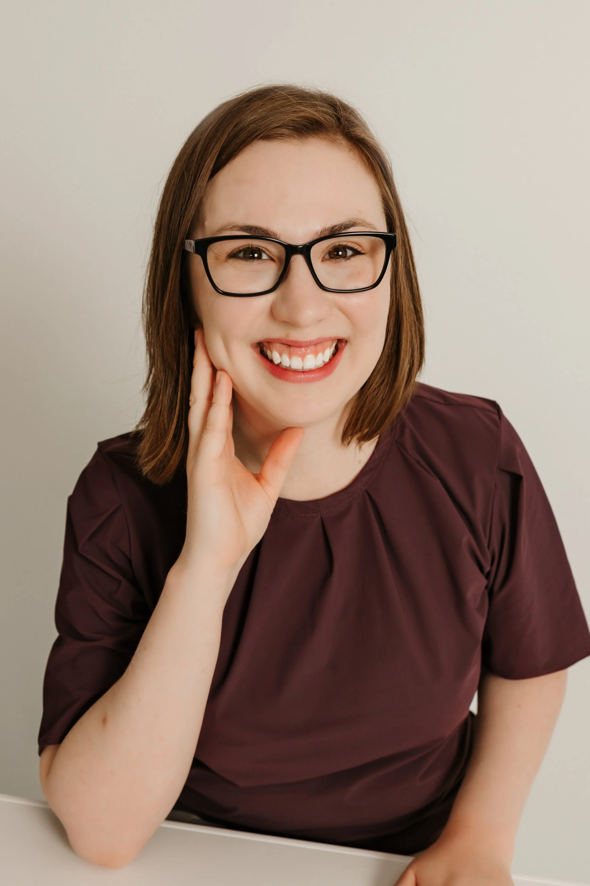 A woman with short brown hair, black glasses, and a maroon shirt, smiling warmly with her hand resting on her cheek.