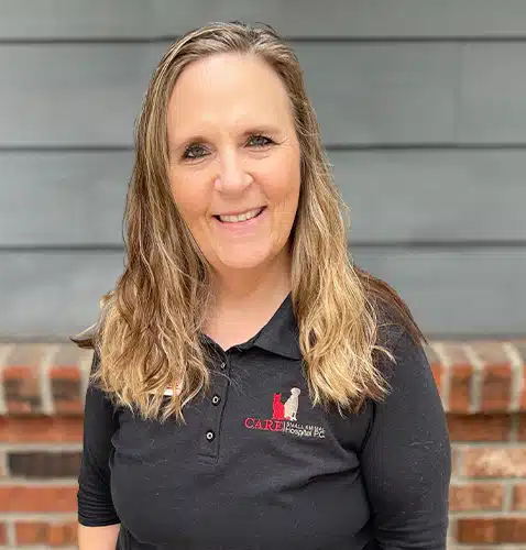Woman with blonde hair smiling, wearing a black shirt with a logo, standing outdoors with a brick wall and gray siding in the background.