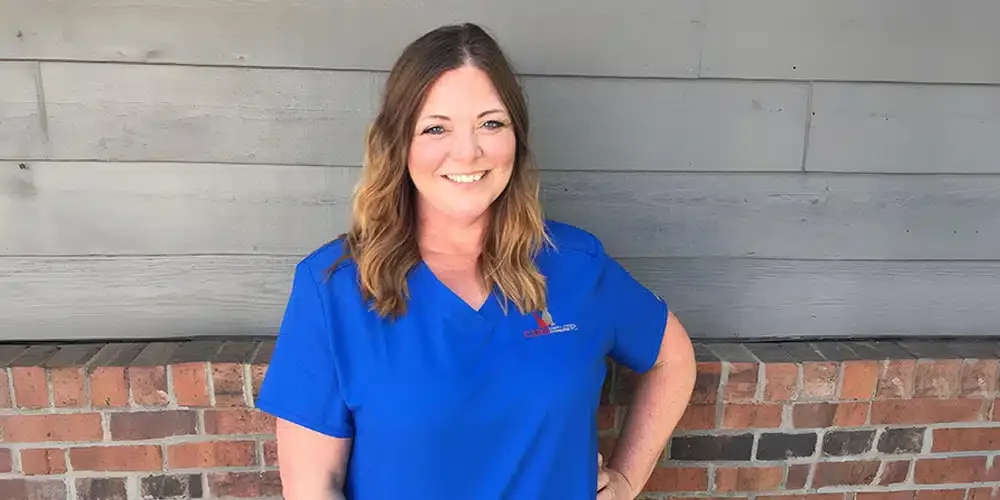 A woman with long, wavy brown hair, wearing a blue medical scrub top, smiling, standing outside in front of a gray wooden wall and a brick ledge.