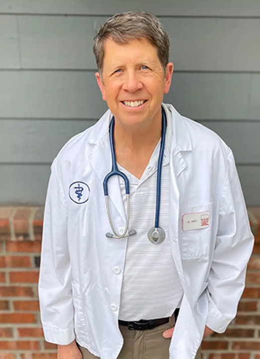 A smiling male healthcare professional wearing a white lab coat and a stethoscope around his neck, standing outdoors in front of a gray siding wall and a brick foundation.