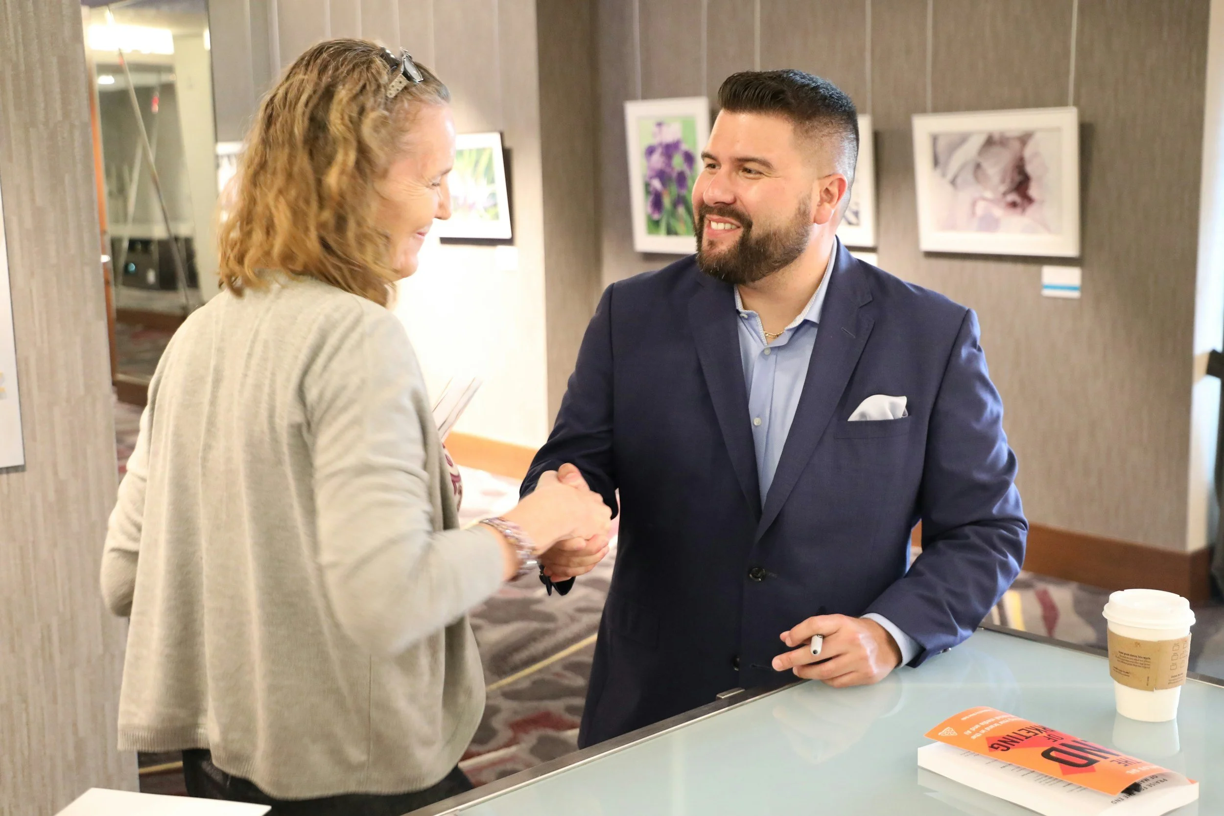 A man in a navy suit and a woman in a beige coat shaking hands inside a gallery or conference space. The woman is smiling and holding a paper, and the man is smiling back with a coffee cup and a magazine on a glass table.